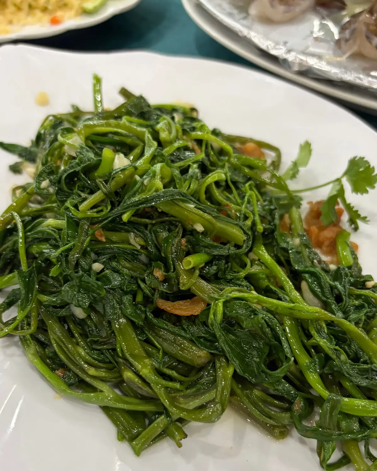 白い長方形の皿に、緑色の細長い野菜がたっぷり盛り付けられています。野菜は柔らかく、細かく刻まれたニンニクや薄く切られた玉ねぎが混ざり、軽く焦げた部分も見えます。右下には少量の赤いソースとパセリの葉が添えられています。
