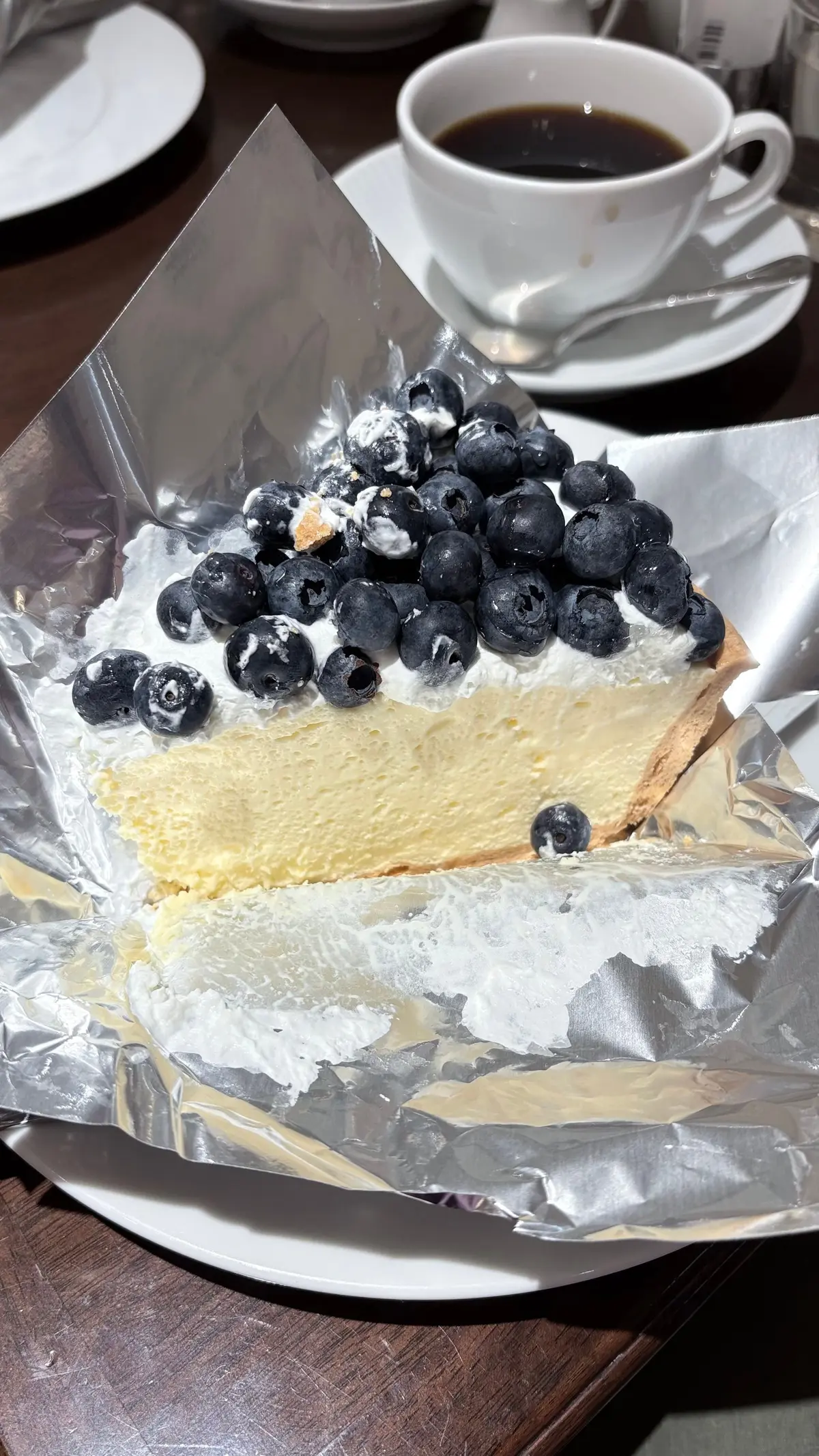 アルミホイルに盛り付けられたチーズケーキ。表面にはホイップクリームとブルーベリーがたっぷり。背景にはコーヒーのカップが置かれている。