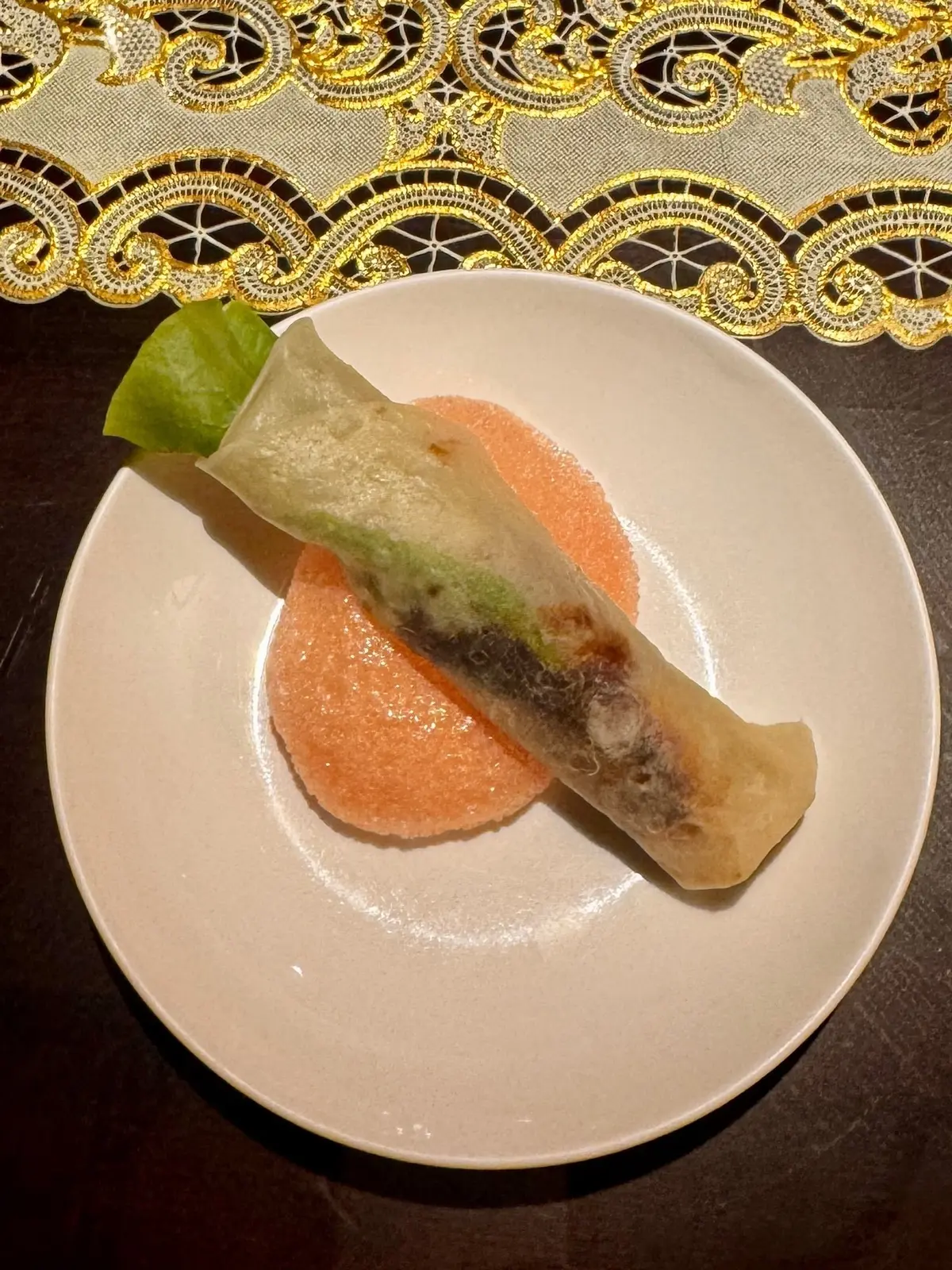 白い皿に、緑色の春巻きとオレンジ色のソース、レタスが盛り付けられている。春巻きは斜めに置かれ、上品なテーブルクロスが背景にある。
