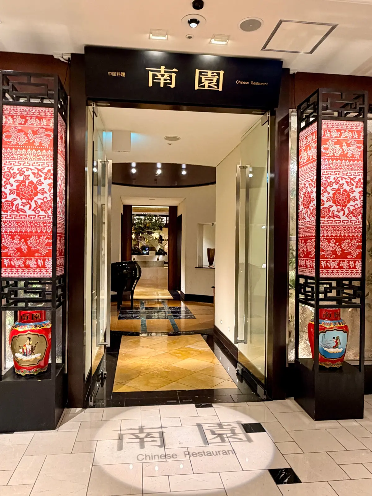 中国料理店のエントランス、ガラスのドアと赤い装飾が施された柱、床はタイルで、店内にはテーブルと花が置かれている