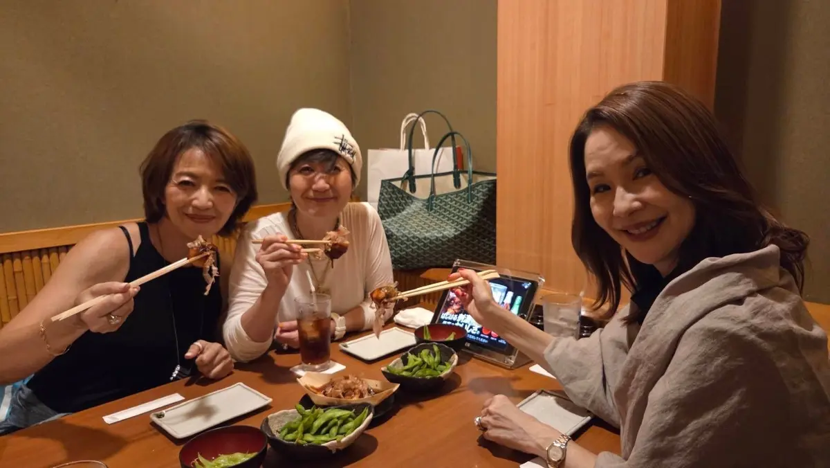 3人の女性が木製のテーブルに座り、料理を楽しんでいる。中央の女性は白い帽子をかぶり、右の女性はタブレットを操作している。各女性は箸で肉料理を食べている。テーブルにはエビ、豆腐、豆、ドリンクが置かれている。