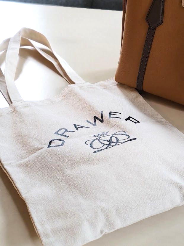白いキャンバストートバッグに、青字で「Drawer」の文字とブランドロゴが刺繍されている。背景には、ベージュ色のレザーバッグが一部写っている。