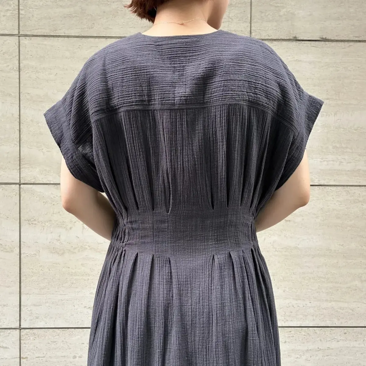濃いグレーのロングワンピース。背中心に縦のプリーツ、ウエスト部分にギャザー。肩はラグラン袖で、ゆったりとしたシルエット。背景はベージュのタイル壁。