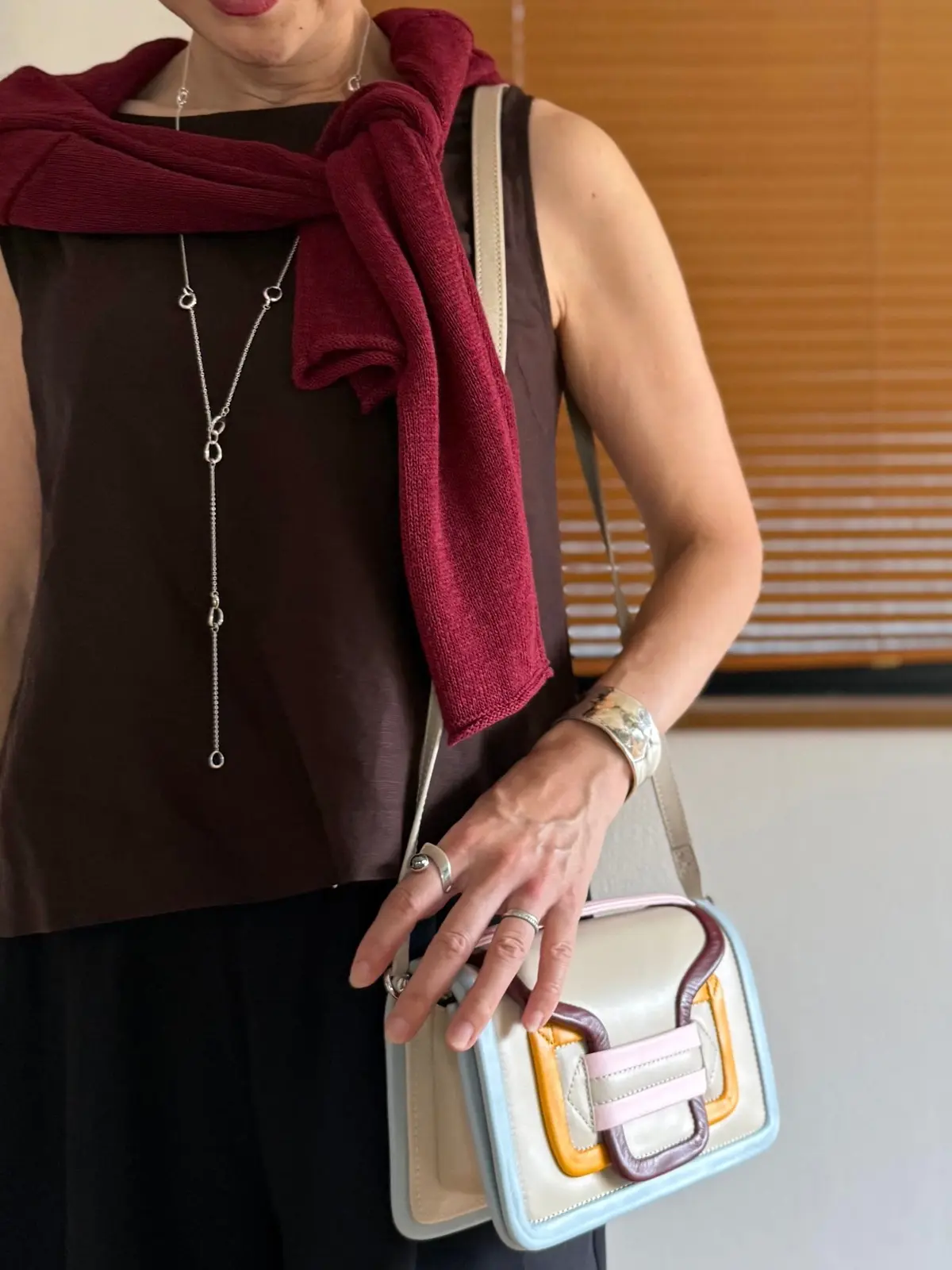 茶色のトップスに赤色のマフラーを巻いた女性。ベージュのバッグを持ち、シルバーのロングネックレスと腕時計を身に着けている。背景にはブラインドが見える。