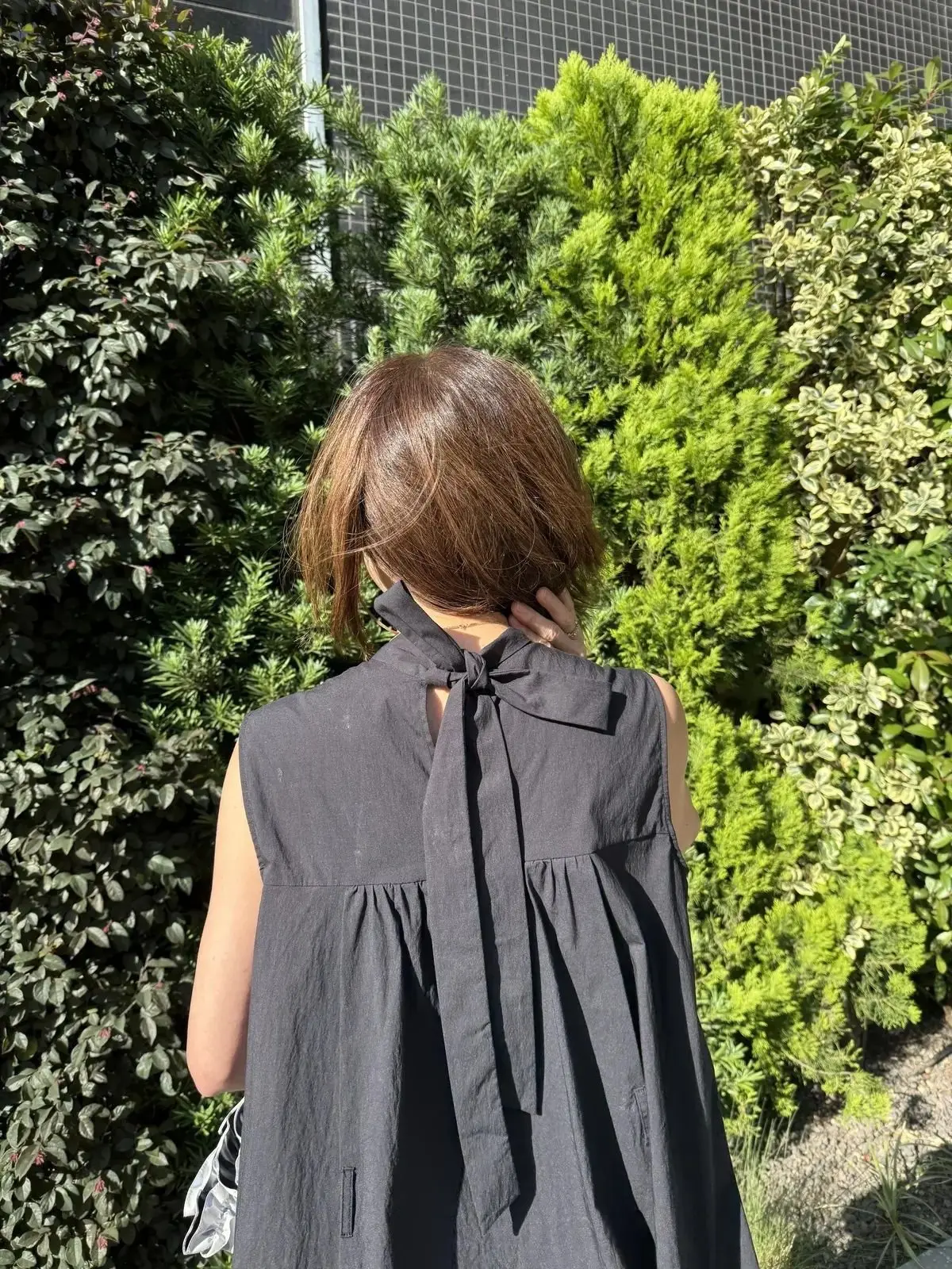 背面から見た女性が、グレー無地でリボン付きの半袖トップスを着て、緑豊かな低木に囲まれた屋外で手で首を触えている