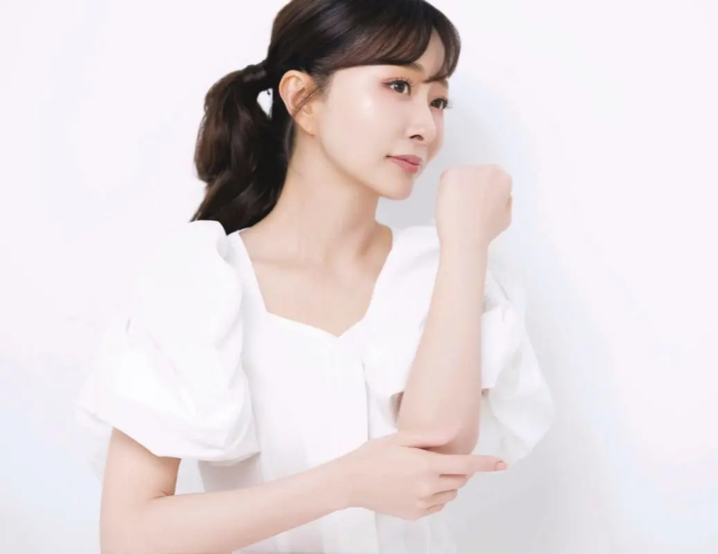 白いブラウスを着た女性が、片腕を曲げて肘のあたりを触っている。茶色の髪を後ろでまとめている。背景は白。