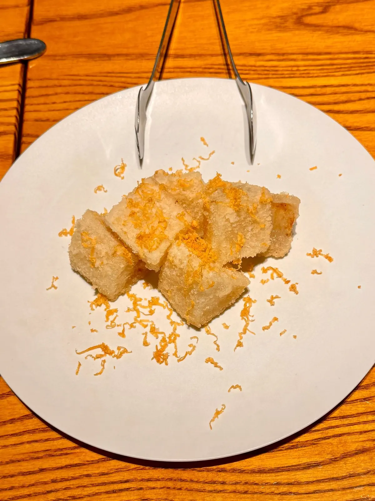 白い皿に、オレンジ色のピールを散らした、四角い揚げ物が3つ置かれている。真上から見た写真で、トングが料理の上に添えられている。