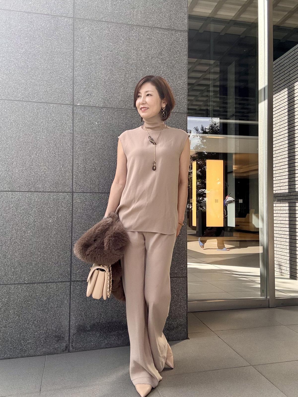ベージュのパンツスーツに、茶色のファーショールを羽織った女性。ネックレスをつけ、ポーズをとって立っている。背景にはガラス張りの建物がある。