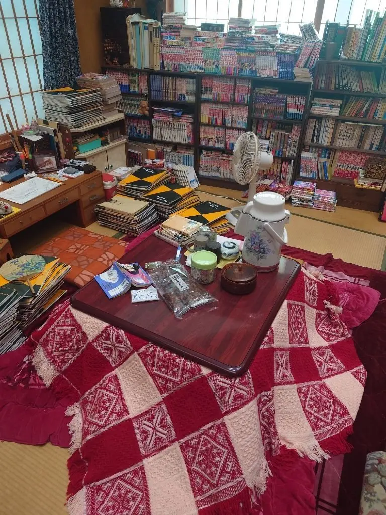畳敷きの部屋で、赤いキルトの上に木製のトレーが置かれ、お茶菓子や茶器が並んでいる。背景には、漫画や書籍がぎっしりと並んだ本棚と、作業机がある。扇風機が置かれ、窓から光が差し込んでいる。