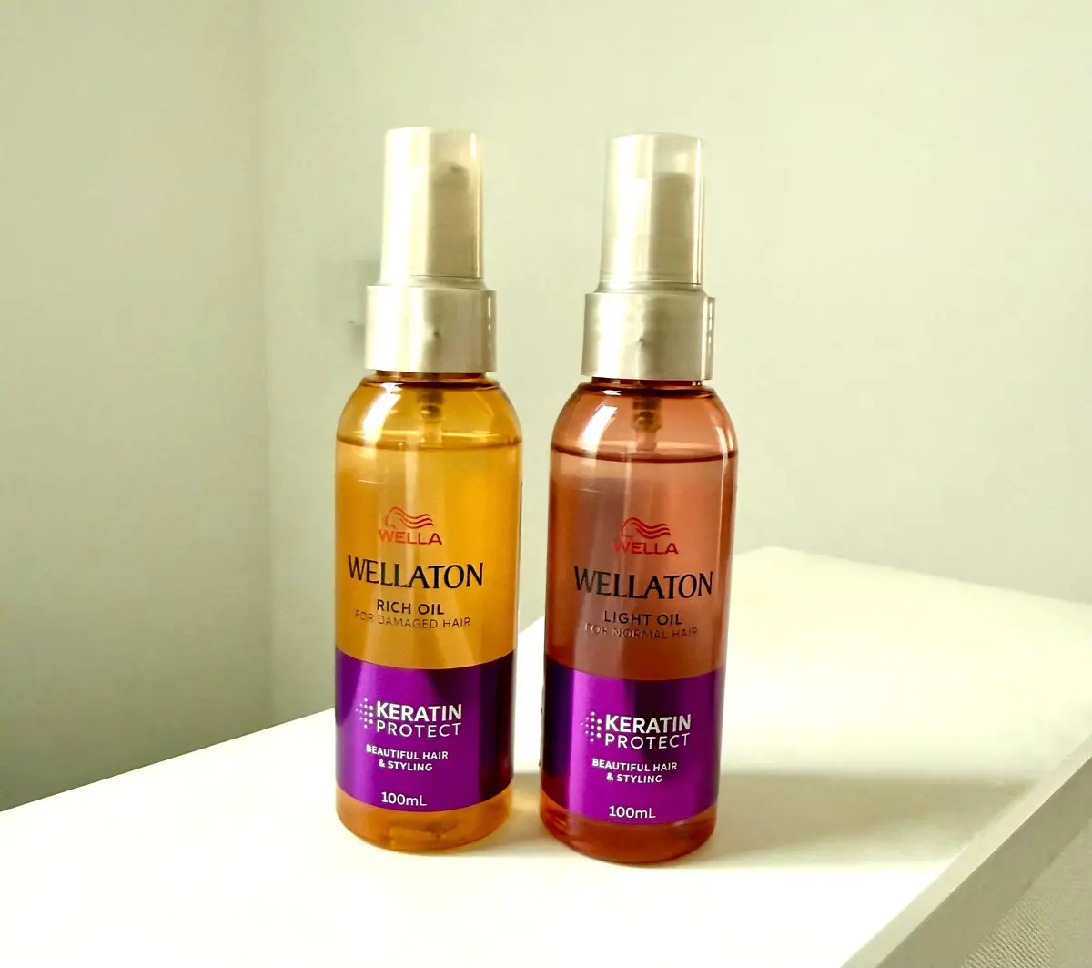 ウェラ（WELLATON）のヘアオイル2本セット。左は「リッチオイル」、右は「ライトオイル」。それぞれオレンジ色のボトルで、100ml。背景には鏡と棚が写っている。