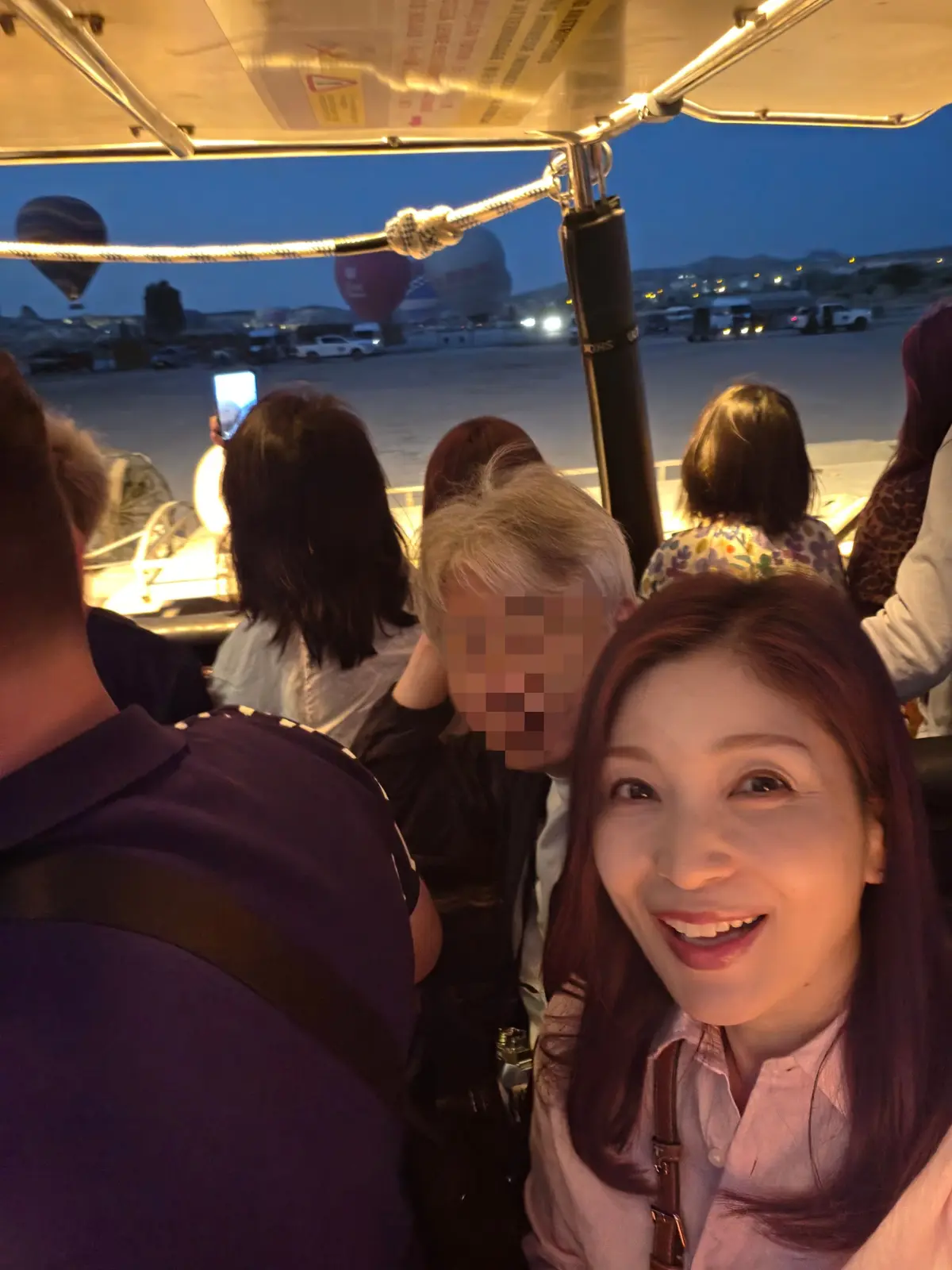 気球が浮かぶ夜空を背景に、ゴンドラ内で笑顔の女性が自撮りをしている。周囲には他の乗客がおり、ゴンドラ内部の構造も見られる。