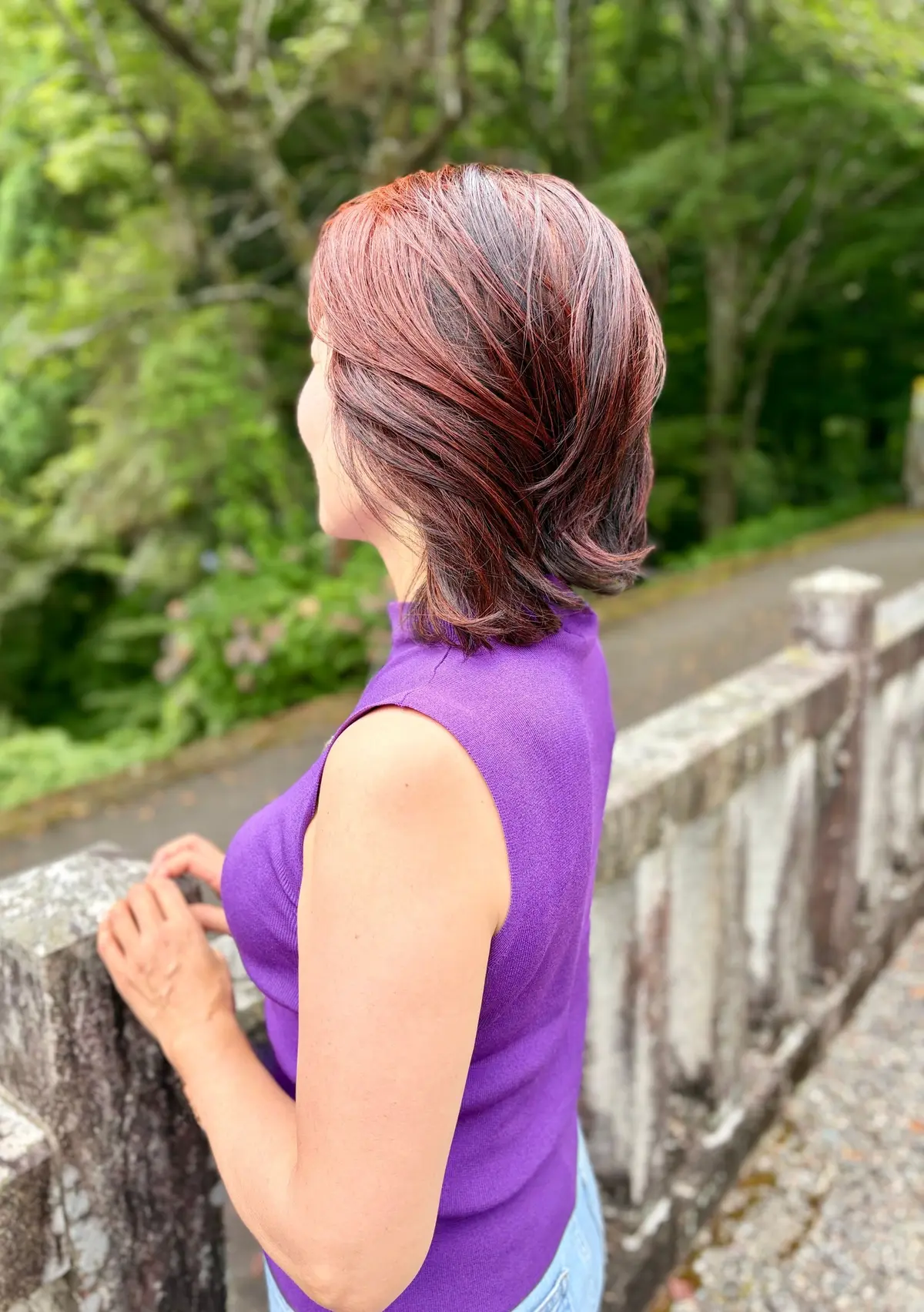 紫色のタンクトップを着た女性が、石の橋の欄干に手をついて、緑豊かな景色を眺めている横からの写真。女性の髪は短く、赤茶色に染まっている。