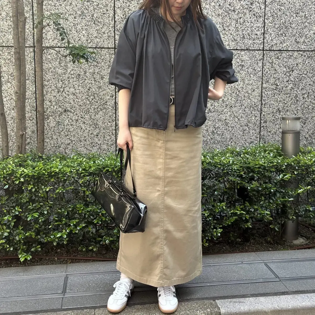 黒のジャケット、ベージュのロングスカート、ホワイトのスニーカーを着た女性が、黒のハンドバッグを左手に持ち、石造りの壁と低木の背景に立っています。