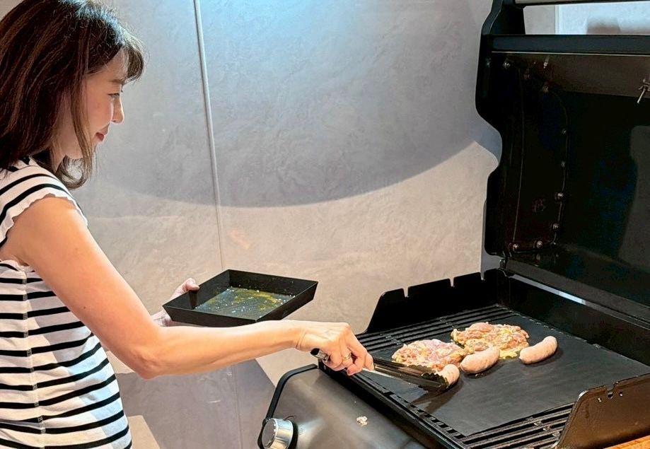 女性が屋内グリルで肉を焼いている。黒と白のボーダーのトップスを着て、フライ返しで肉を返し、別の容器でソースを注いでいる。背景には白い壁と、グリル内部が見える。