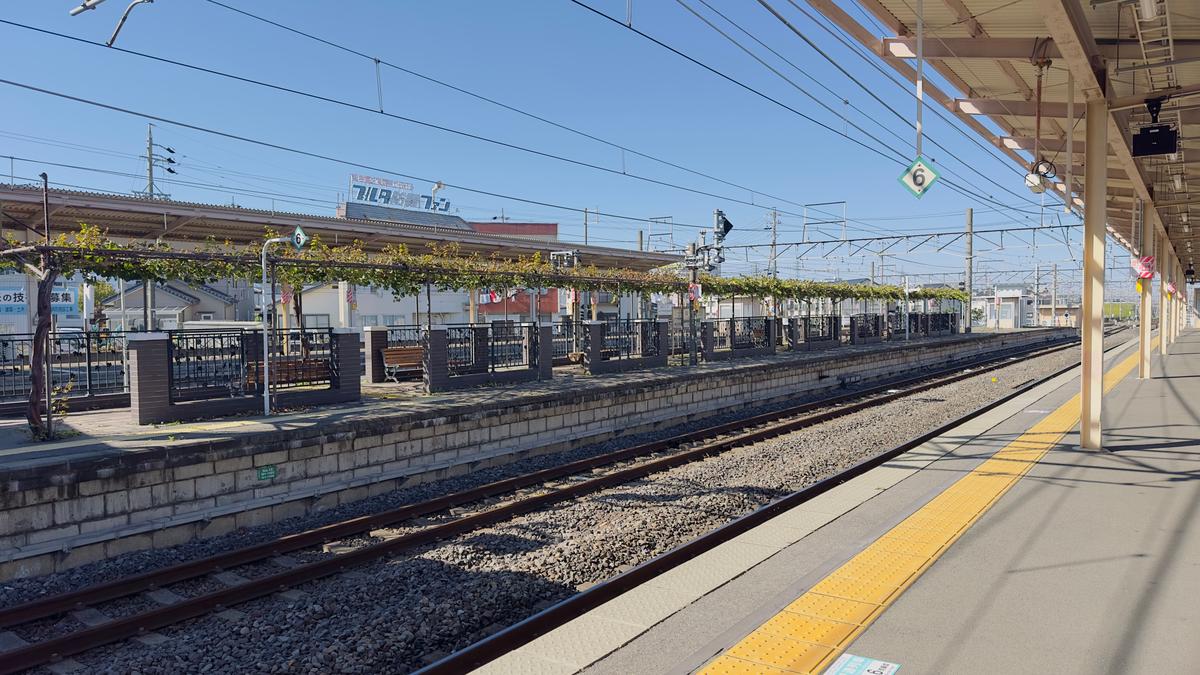 晴天の日の駅のホーム。線路脇には緑の植物が茂り、遠くに建物の看板が見える。ホームには黄色いラインが引かれ、プラットフォームの屋根が上空に伸びている。