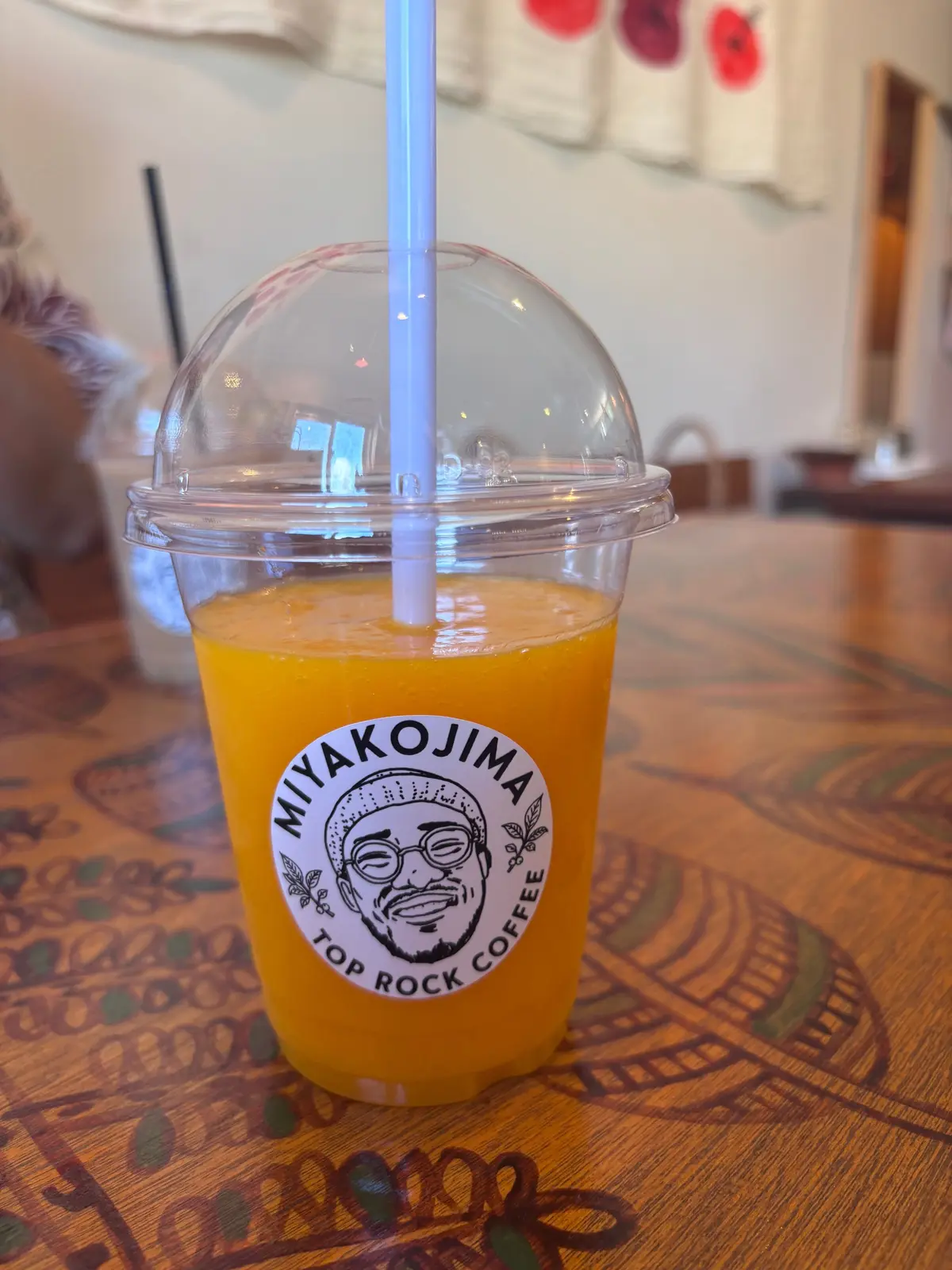 透明なプラスチックカップに入ったオレンジ色のドリンク。白いストローが挿入され、カップには「MIYAKOJIMA TOP ROCK COFFEE」のロゴがデザインされている。