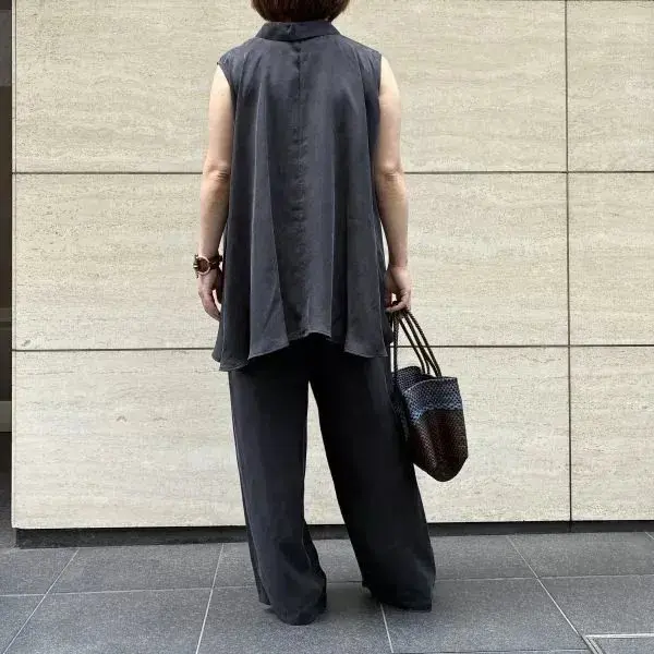 黒いスリーブレスのシャツとワイドパンツを着用した女性が、石の壁の前に立っている。右手に編み込みのバッグを持っている。
