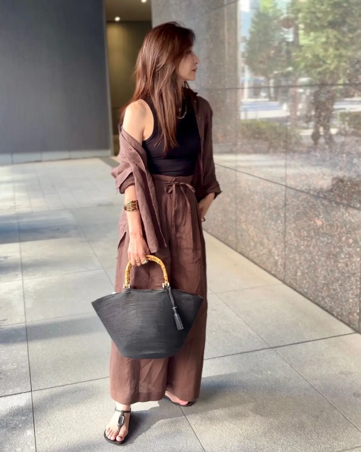 茶色のワイドパンツとタンクトップに、同系色のシャツを羽織った女性。黒いサンダルを履き、黒いバッグを持つ。背景はモダンな建物の壁。