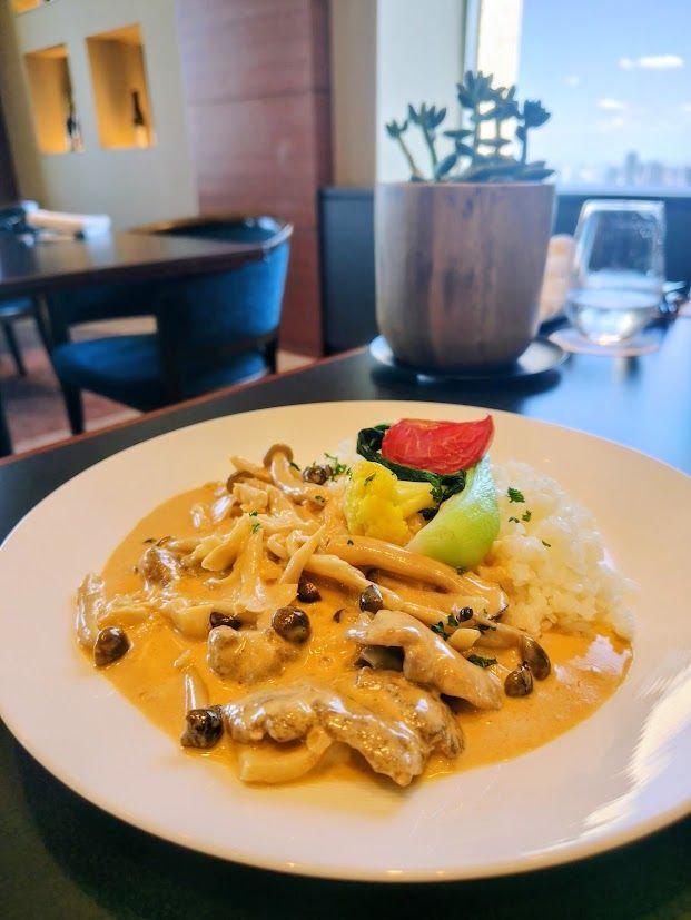 白い皿に盛り付けられた、クリーム色のソースとキノコ、肉の料理。右側に白米、レモン、トマト、葉野菜の飾り付け。背景には観葉植物と窓からの景色。
