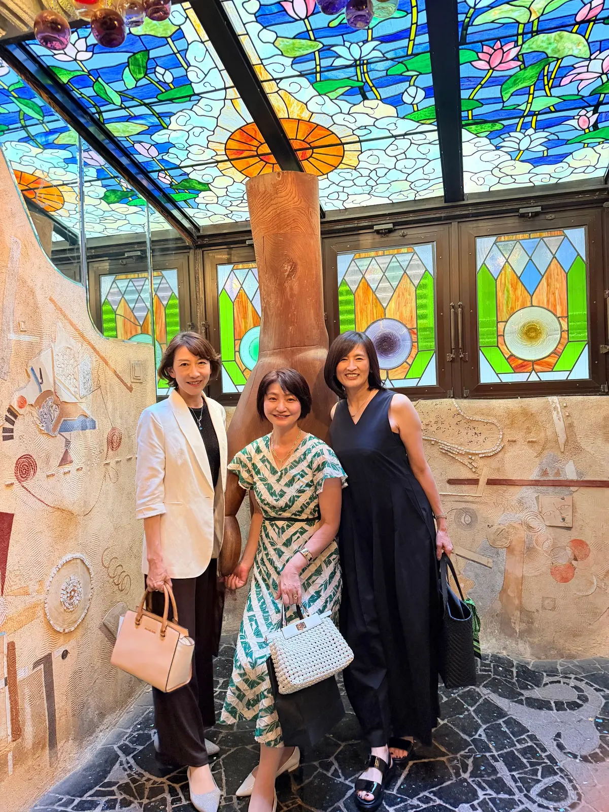 ステンドグラスの天井の下、3人の女性が笑顔で並んで立っている。左から白のジャケット、中央は緑色の花柄ワンピース、右は黒のワンピース。背景には、ベージュの壁に模様が描かれ、床は黒と白のモザイク模様。