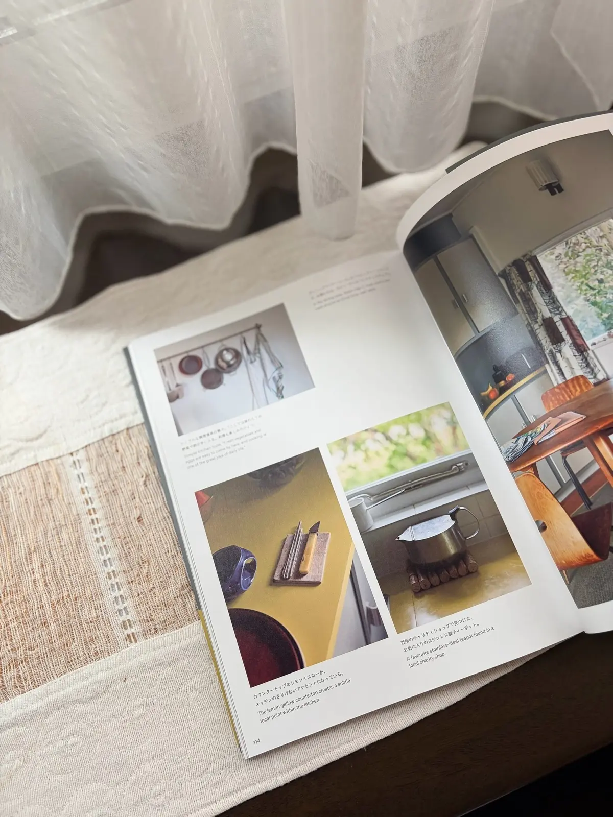 開いた雑誌の上に置かれた白いソファ。雑誌には、キッチンやリビングのインテリア写真が掲載されており、木製の家具や自然光が特徴。右ページには、レトロなデザインのテーブルと椅子が写っている。