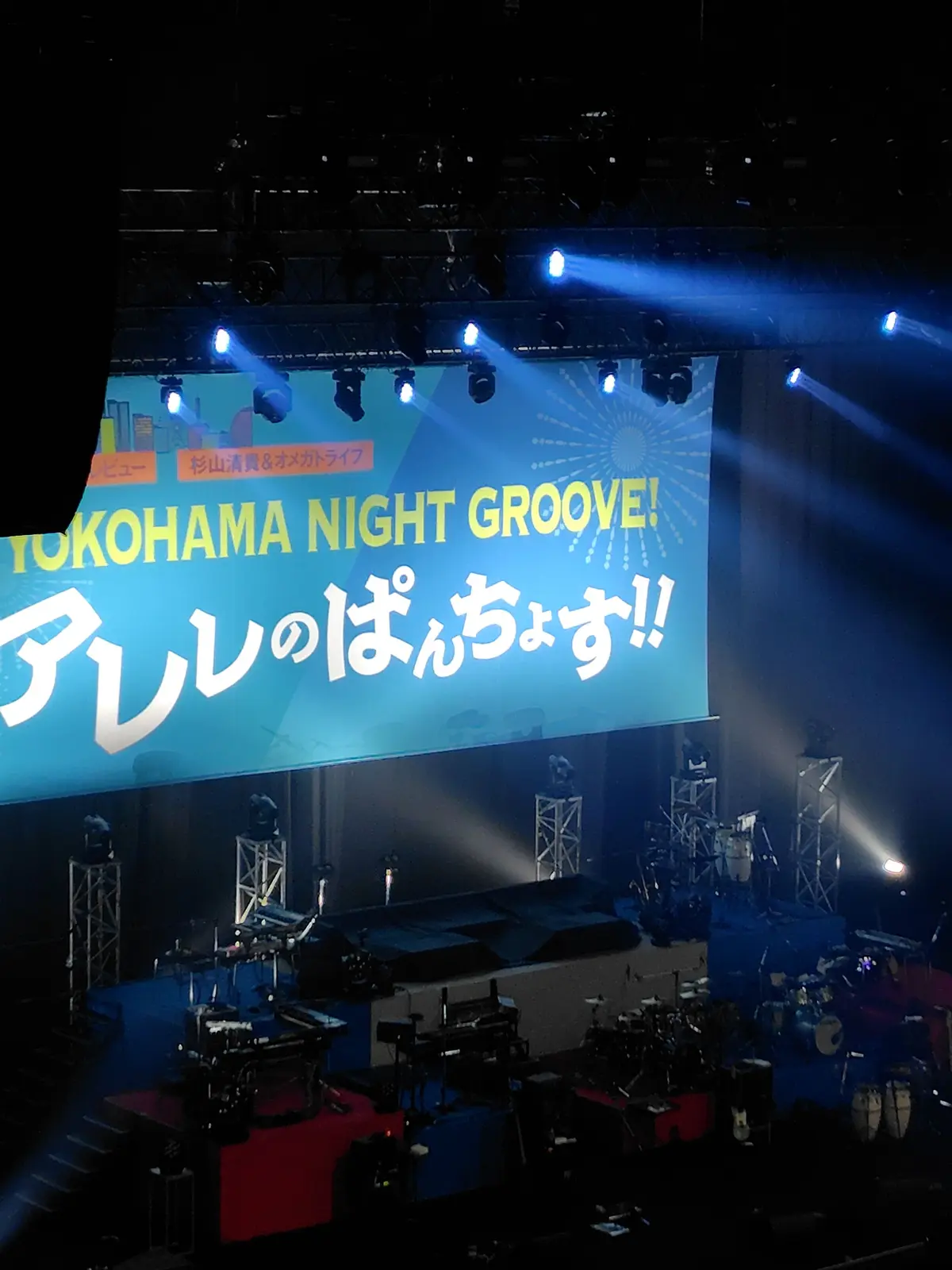 横浜ナイトグルーブのライブステージ、青い背景に白い文字で「アツいねんじゃす!」と表示され、照明がスポットライトで照らしている