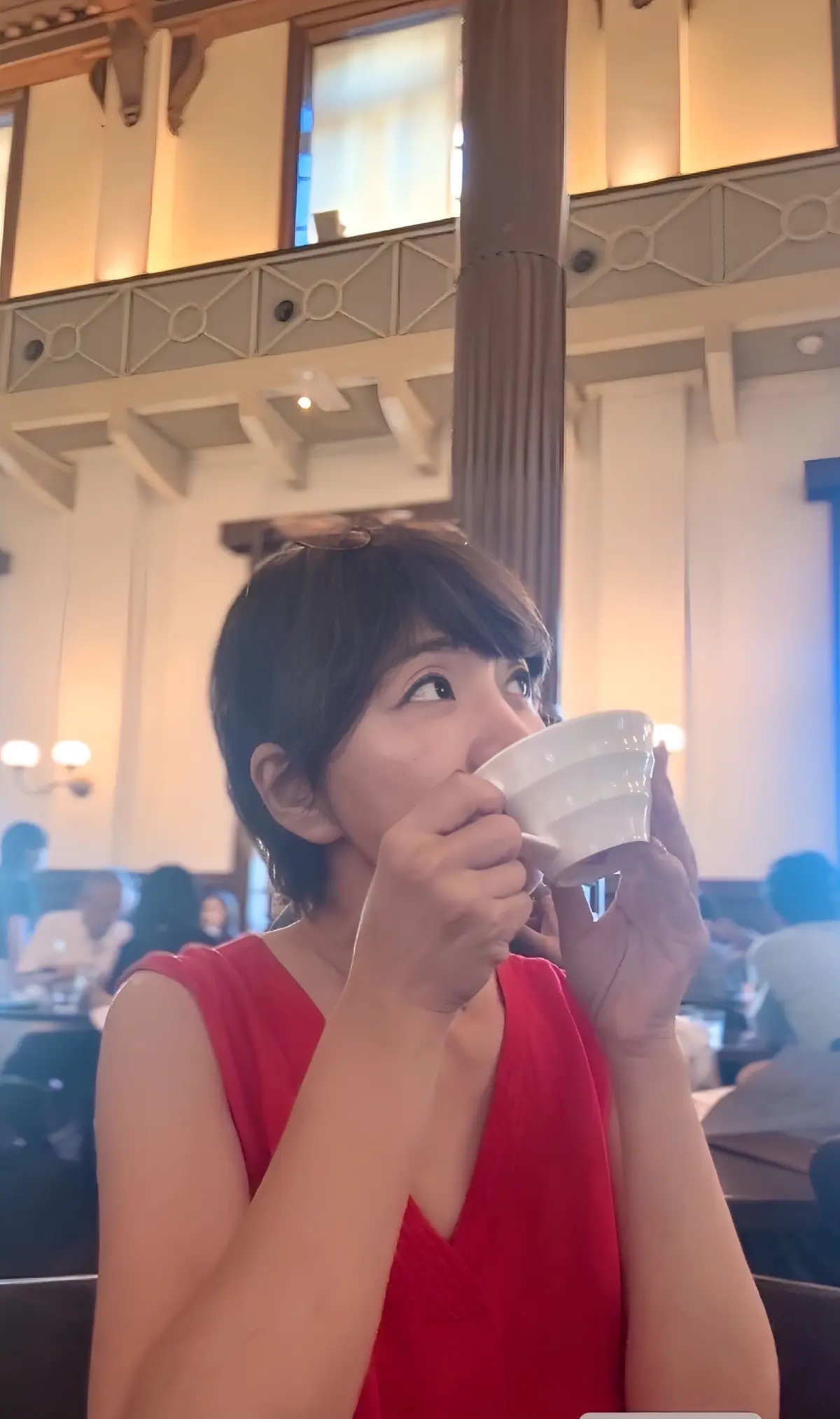 赤いノースリーブの女性が、白いカップに入った飲み物を飲んでいる。背景には、窓と装飾的な柱が見えるレストランの様子。