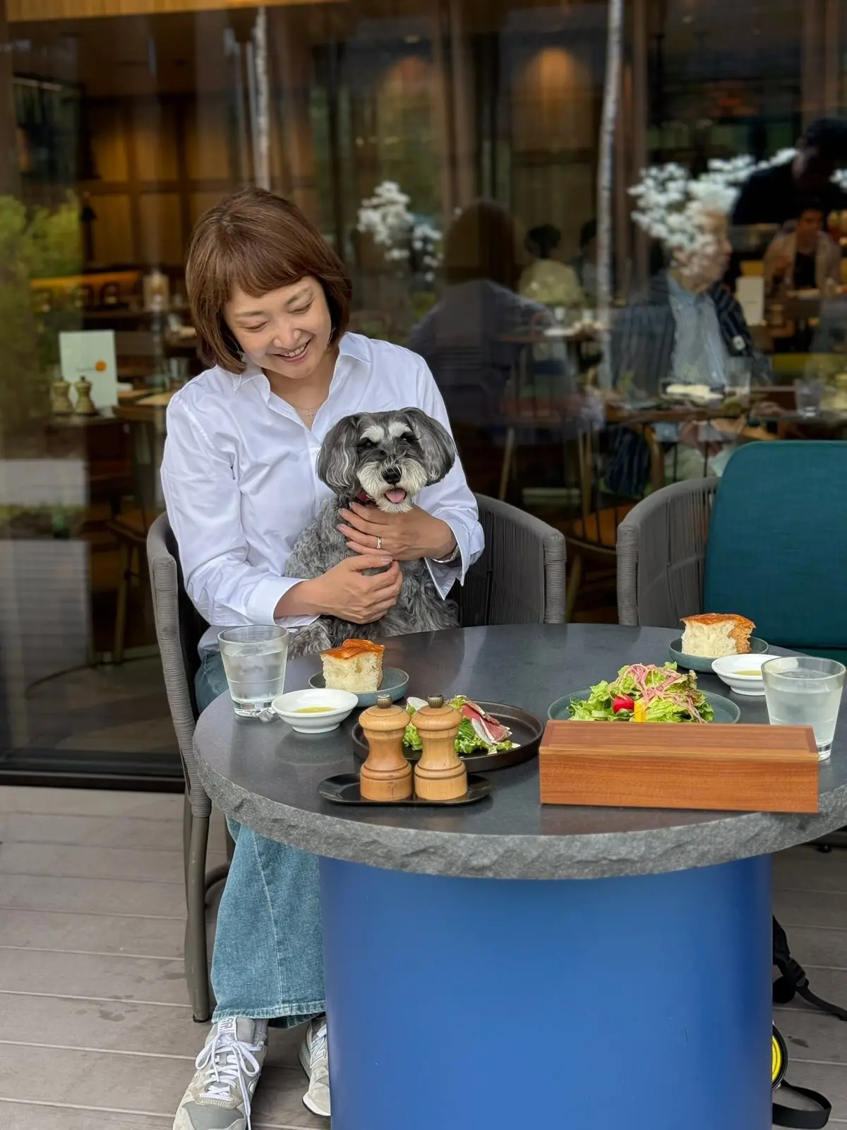 女性がカフェの屋外テーブルに座り、小型犬を抱えています。テーブルにはサラダ、パン、調味料、飲み物が置かれています。