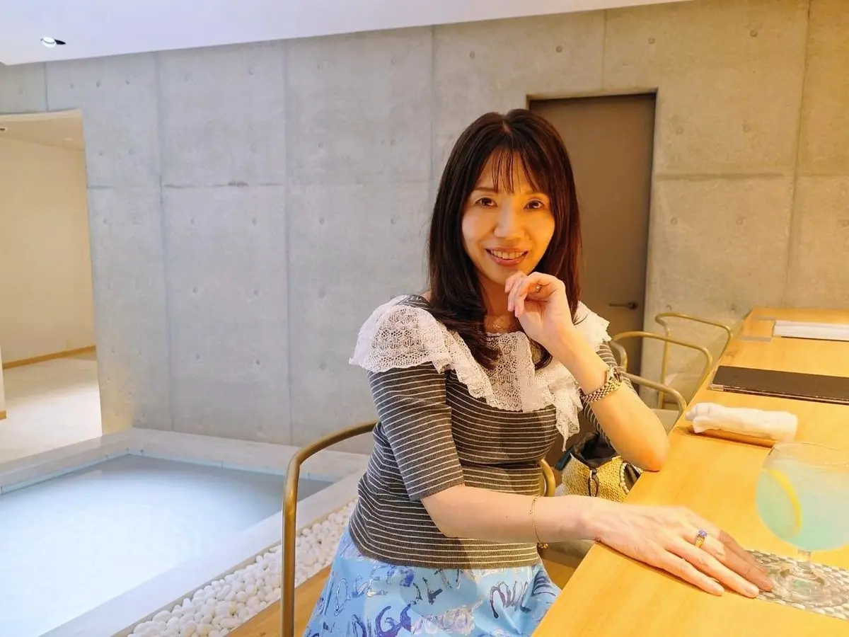 三輪湯河原のバー。笑顔の女性が椅子に座り、テーブルに片手を置いている。青と白のボーダーのトップスに、白いレースの襟が付いている。