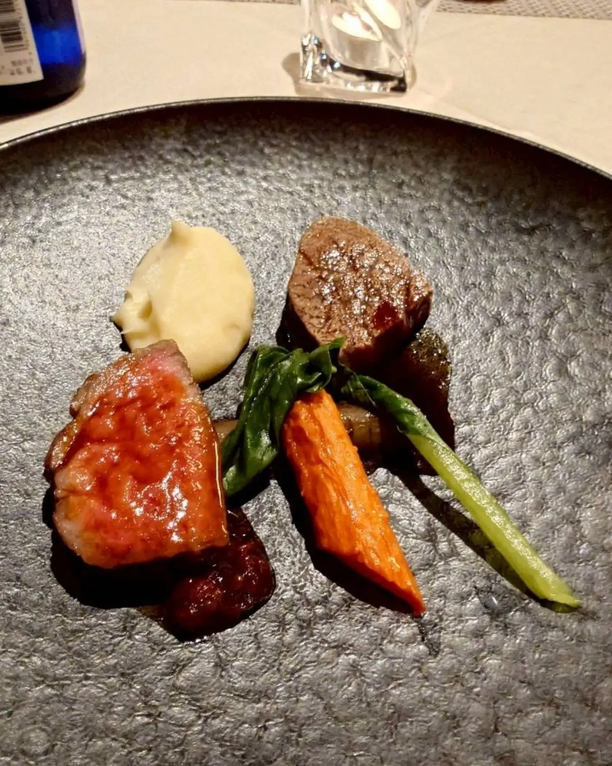 黒い皿に盛り付けられた、牛肉と野菜の付け合わせ。牛肉は赤身と焼き色が付き、マッシュポテト、オレンジ色の人参、緑色のキュウリ、葉物野菜が添えられている。
