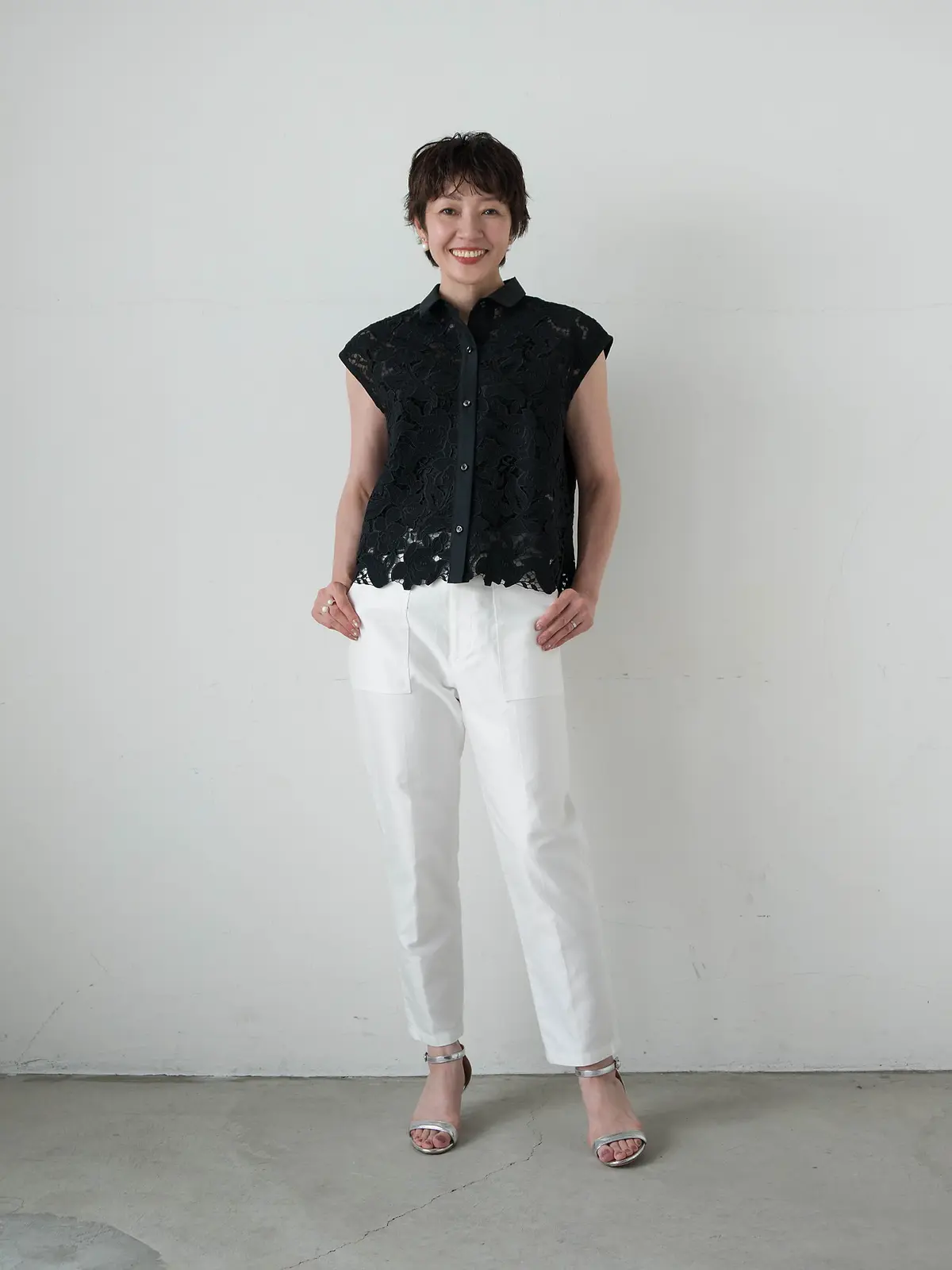 白い無地背景に、黒いレーストップと白いワイドパンツを着た女性が微笑んでいる。足元にはシルバーのサンダルを履いている。