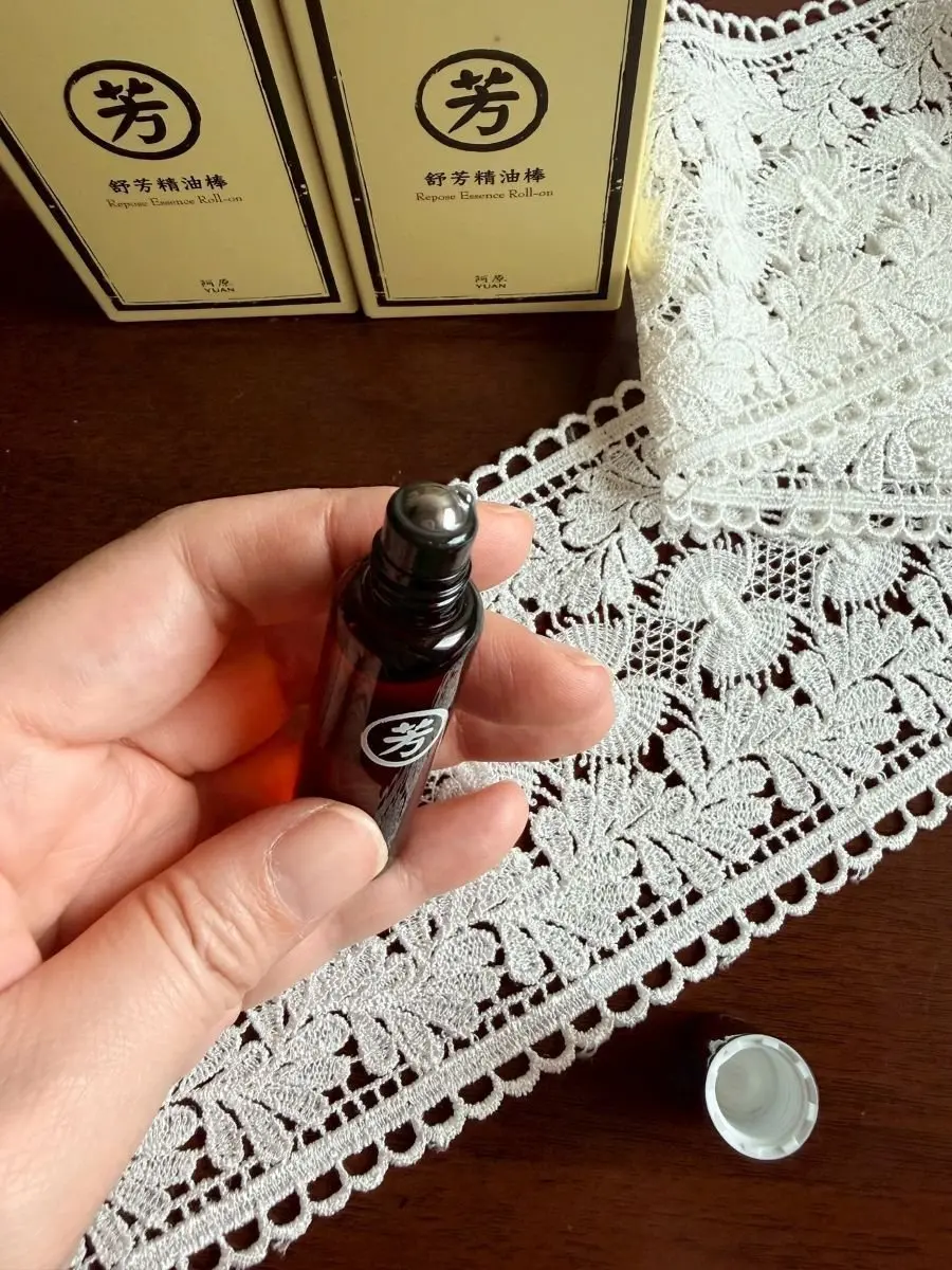 手は茶色のボトル「芳」を持ち、白色レースのテーブルクロス上に置かれた同様のボトルの箱が背景にあり、ボトルのキャップがテーブルの上に転がっている