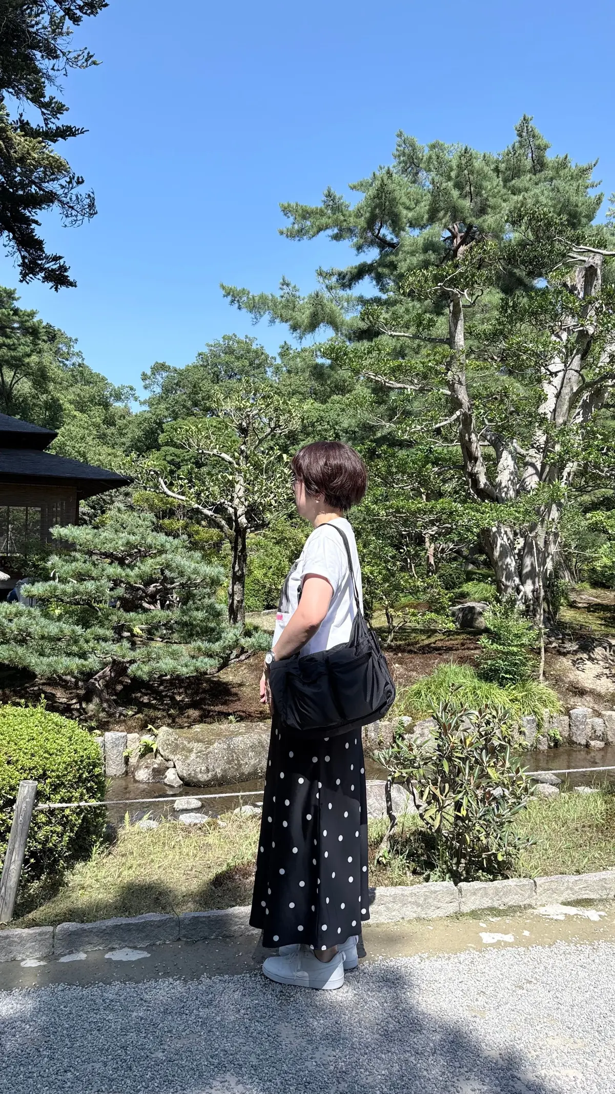 黒地に白いドット柄のロングスカートと白いTシャツを着た女性が、庭園を眺めている。茶色のバッグを肩にかけ、白いスニーカーを履いている。背景には緑豊かな木々と、石で組まれた小道が見える。