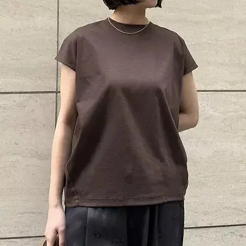ブラウンのカットソーを着用した女性。丸みのある肩と、短めの袖が特徴。シンプルなクルーネックで、細めのシルバーネックレスを着用。背景はグレーのタイル壁。