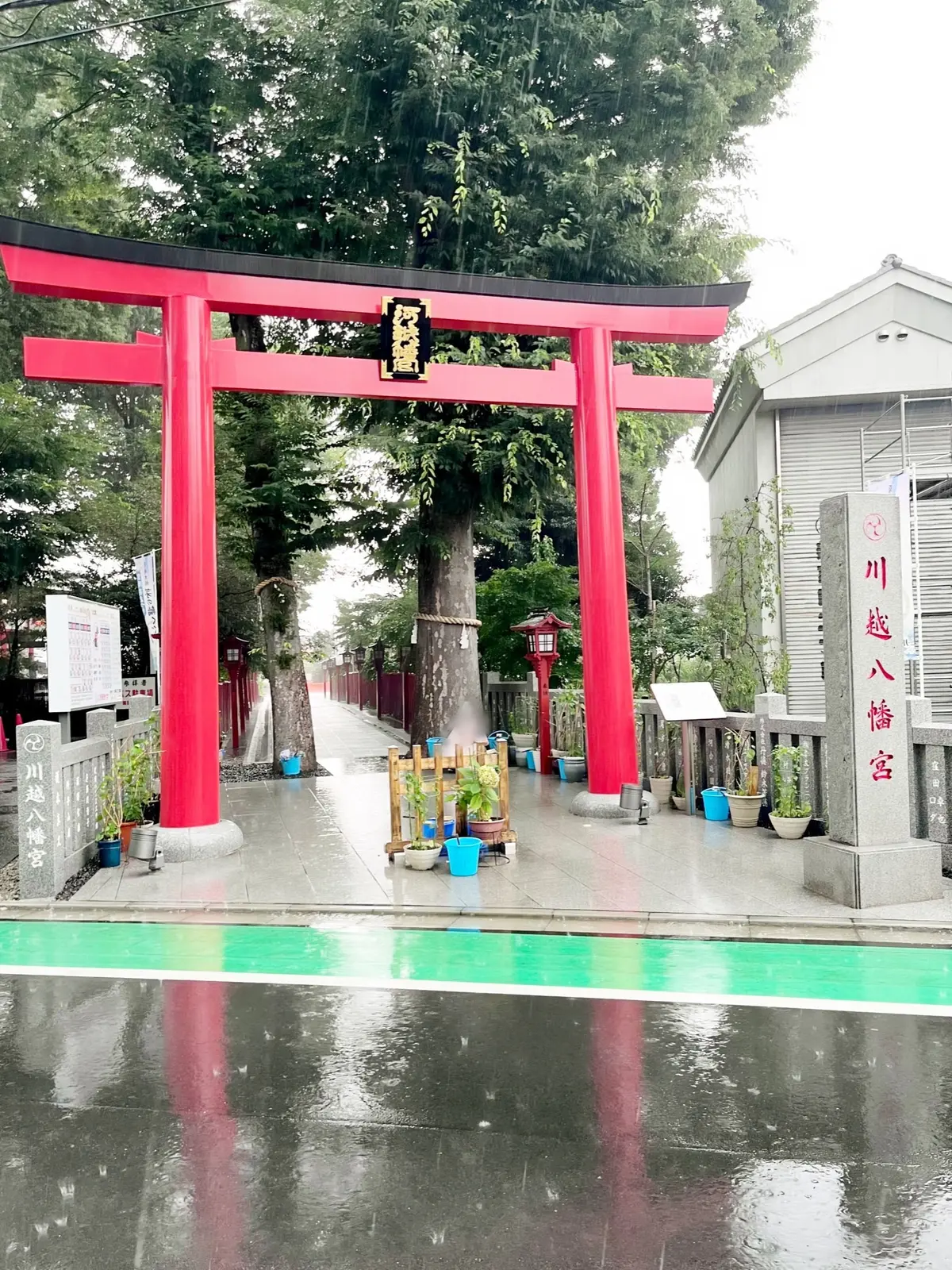 赤い鳥居が緑の木々を背景に立ち、雨の降った路地に石の看板や植木が並ぶ神社入口