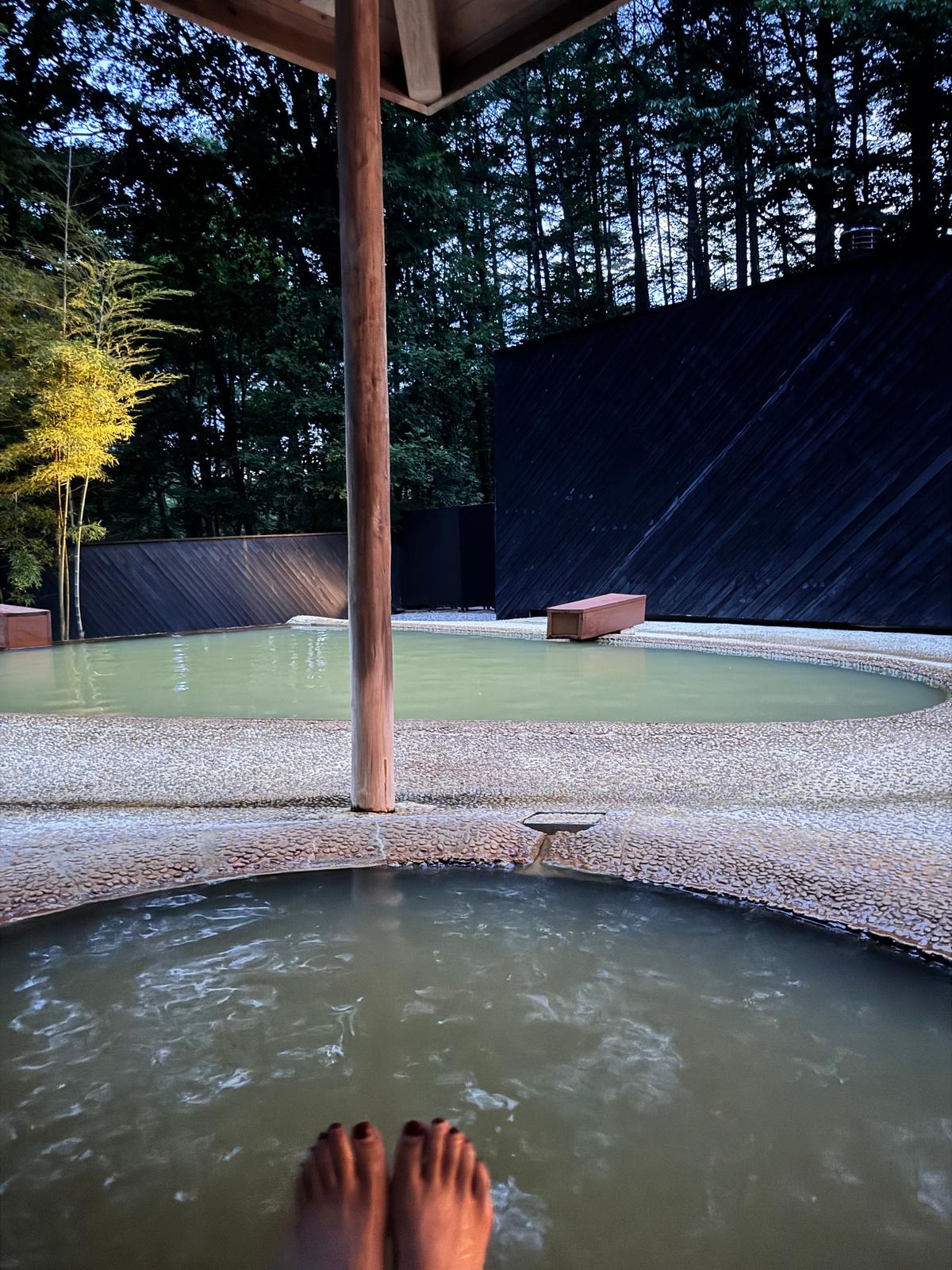 丸い露天風呂に足湯を楽しみ、背景には黒い壁と緑豊かな木々が見える庭園。中央には柱があり、白い砂利が敷き詰められている。
