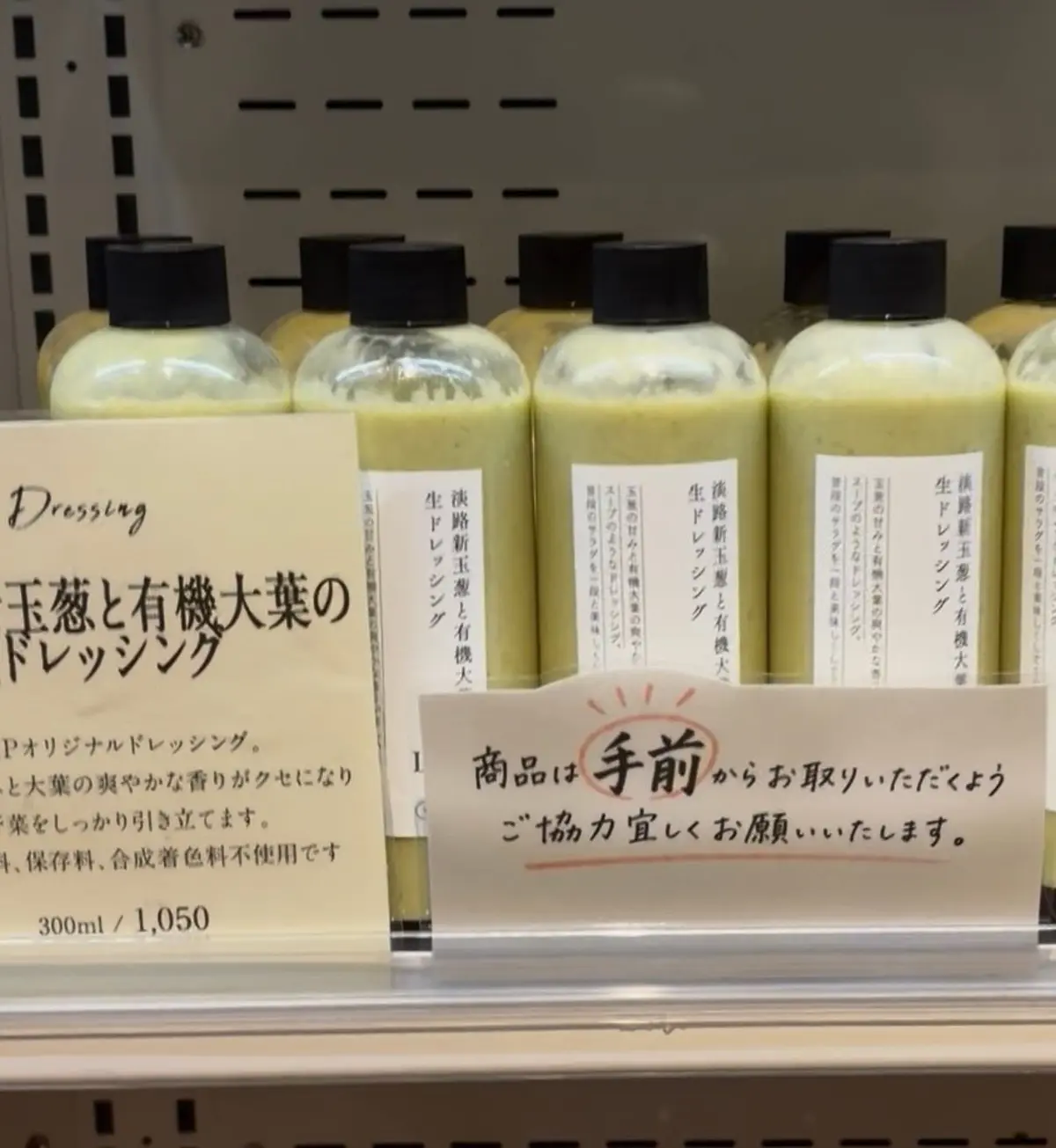 淡色系のボトルに入ったドレッシングが並んでいる。前面には「Dressings 基礎と有機大葉のドレッシング」と書かれたラベルが付いており、価格は390mlで1,050円。手前に「商品は手前でお取りいただきます」と書かれたカードが置かれている。