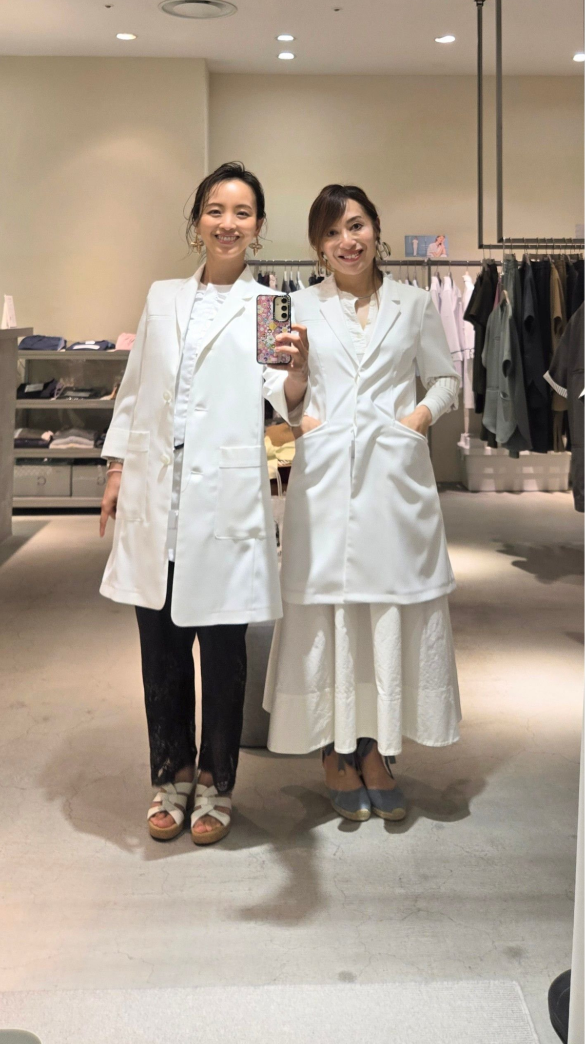 白いコートを着た2人の女性が、明るい衣料品店で並んで立ち、笑顔でセルフ写真を撮っている。左の女性は黒のズボンにサンダル、右の女性は白のワイドパンツにヒール。背景には衣服が並ぶラックと棚があり、店内は清潔感のあるデザイン。