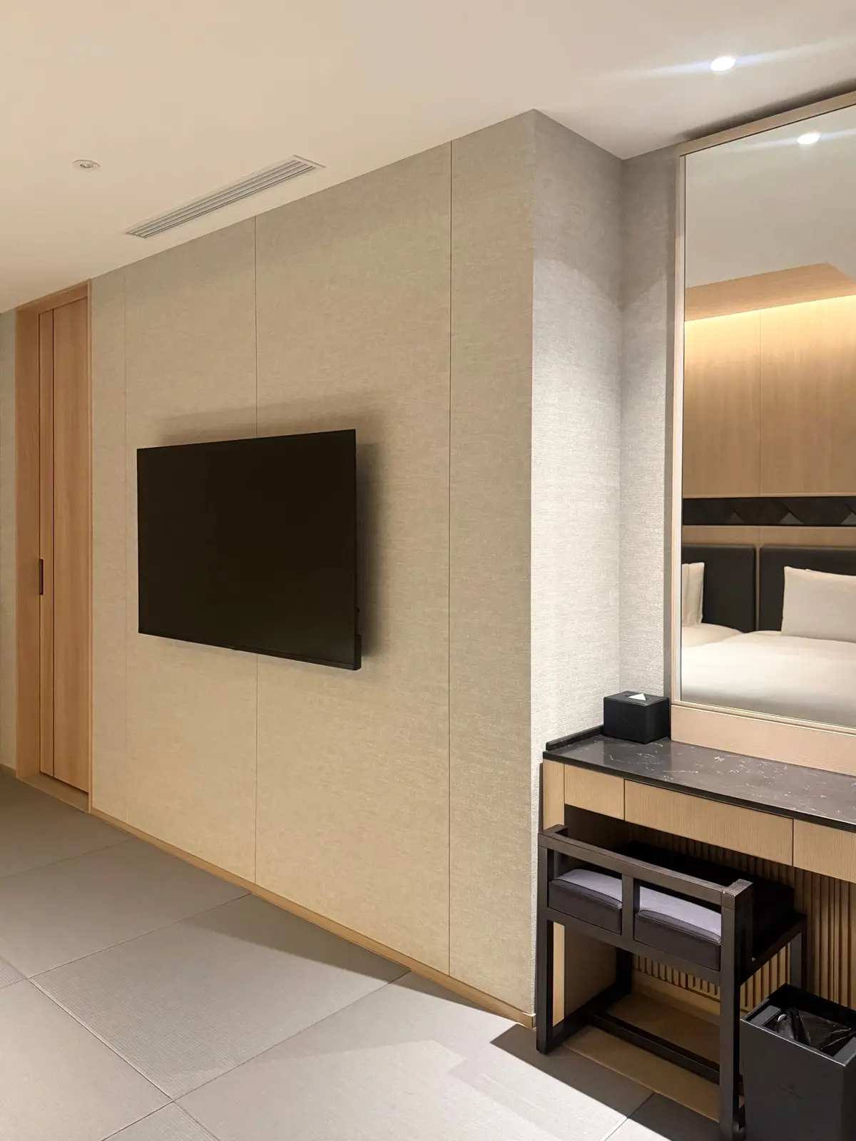 ベージュの壁にフラットテレビが設置された簡潔な部屋で、鏡付きの木製ドレッサーとベンチがあり、ベッドの反射が鏡に映っている