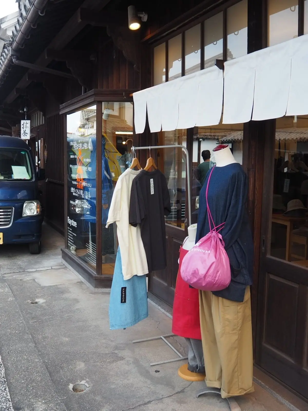古民家風の店先。2体のマネキンが洋服を着て展示されている。左は白と黒のTシャツに青いスカート、右は濃い青のシャツにベージュのズボン。右のマネキンはピンク色のバッグを斜め掛けしている。店の前には紺色の車が停車している。