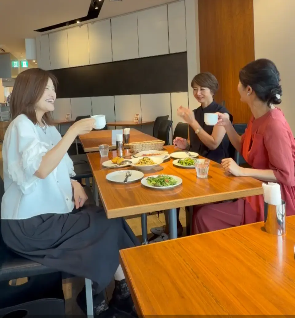 テーブルを囲んで食事をする4人の女性。左から2人目の女性が笑顔で話している。テーブルには料理と飲み物が並び、背景にはエアコンや照明が見える。