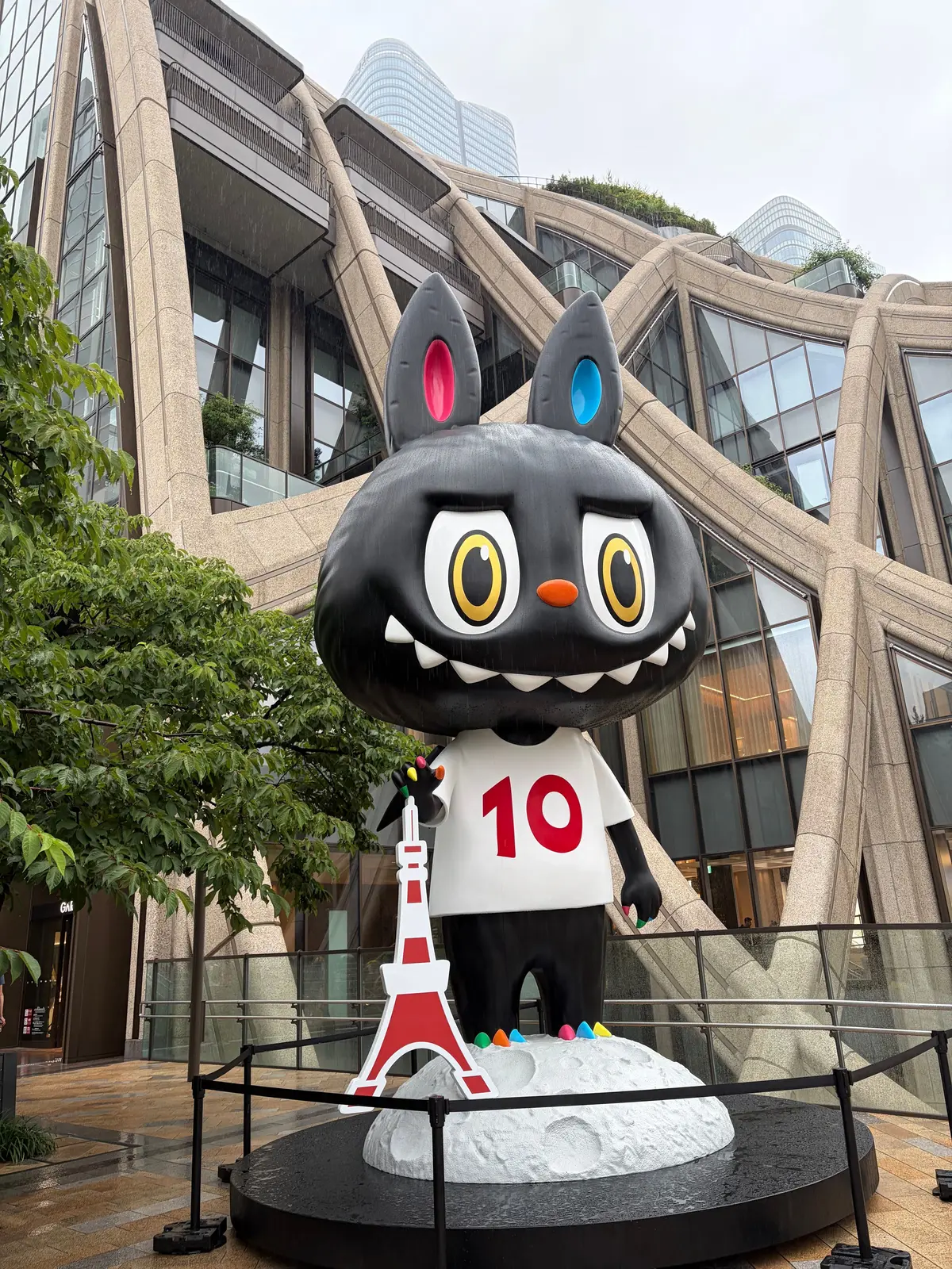 黒猫の巨大なキャラクターが、10と書かれた白いTシャツを着て、エッフェル塔のミニチュアを手に持つ。背景には近代的なガラスのビルが並ぶ。