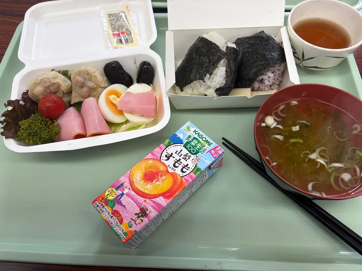 白いトレイに、卵、サラダ、肉、野菜、餃子、ワカメご飯、茶碗、うどん、スープ、果物ジュースの箱が並んでいる