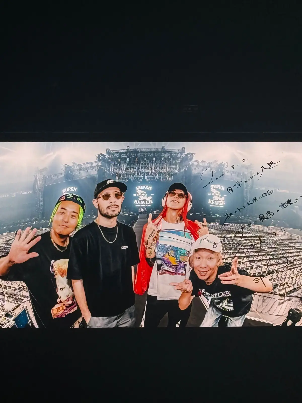 4人の男性がポーズを取る写真。背景には観客席が広がるスタジアムの風景。左から緑色の帽子を被った男性、サングラス姿の男性、赤いゴーグルをかけた男性、白い帽子を被った男性。それぞれが個性的なファッションで、楽しげな表情を浮かべている。