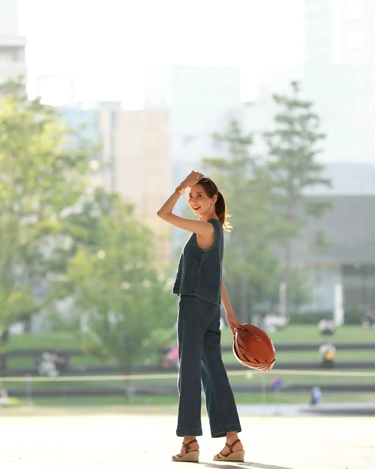 青いデニムのトップスとパンツを着用した女性が、公園で後ろを振り返り笑顔。右手に茶色の帽子を持ち、茶色のサンダルを履いている。背景には緑豊かな木々と建物が見える。