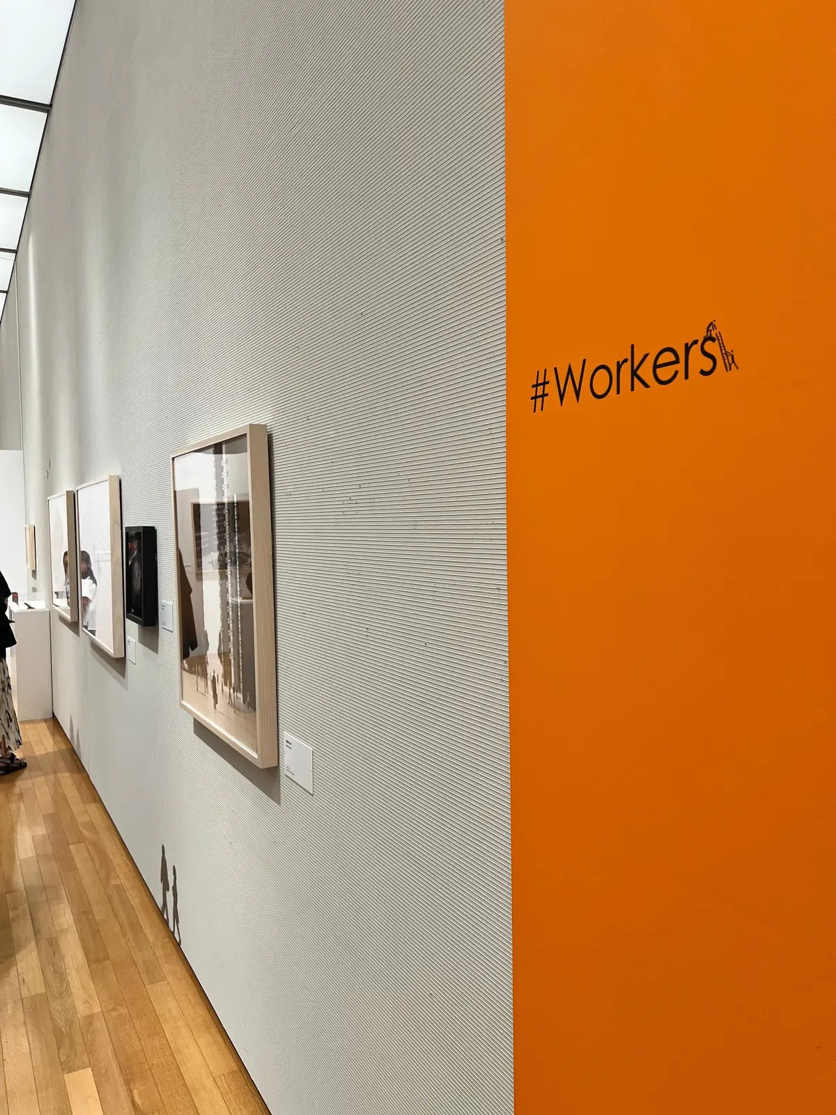 美術館の展示室。左壁には複数の写真作品が額装されて飾られ、右壁は鮮やかなオレンジ色。壁に「#Workers」の文字が白で表記されている。床は木製で、奥には来場者がいる。