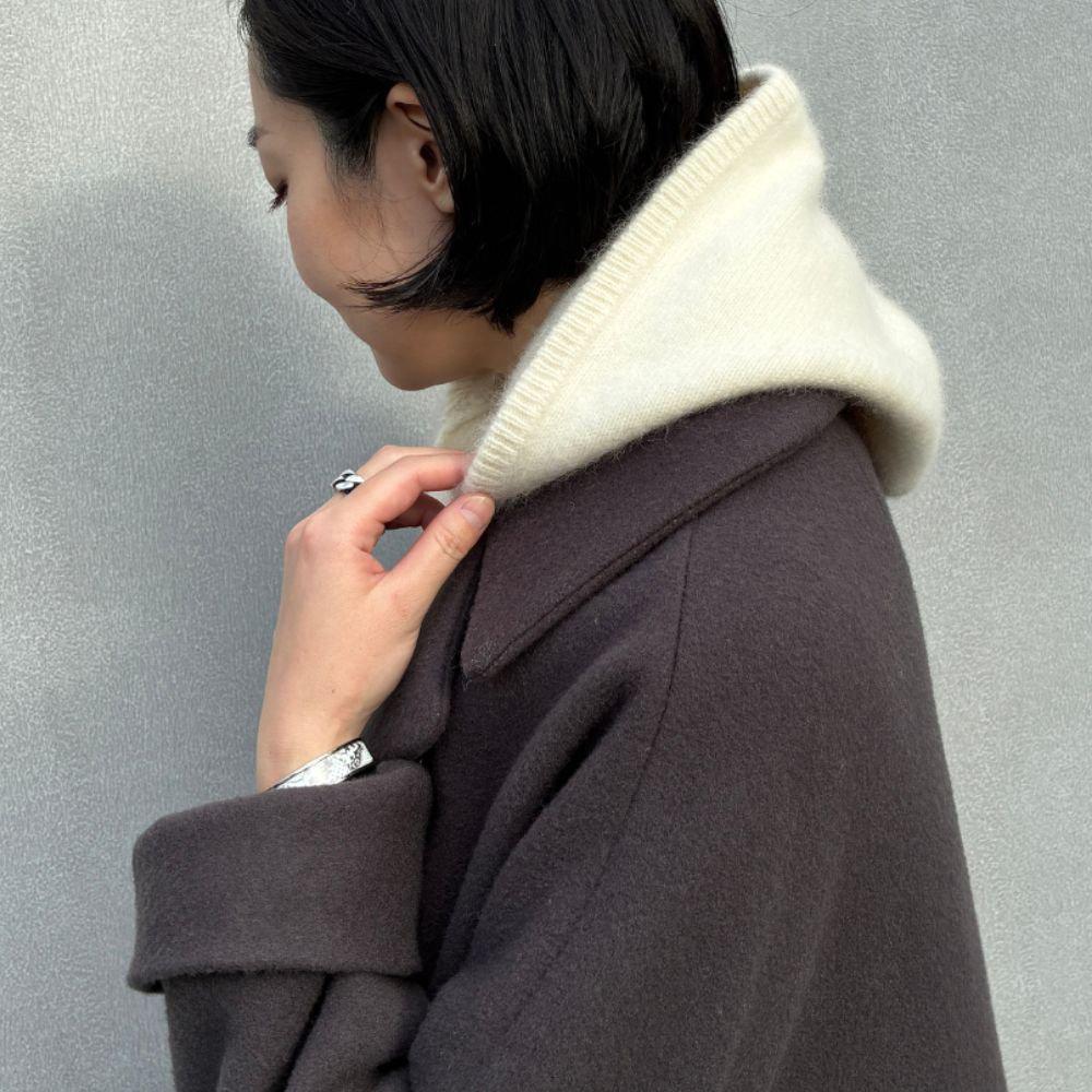 黒髪の女性が、茶色のシンプルなコートと白いタートルネックセーターを着用。右手をコートの襟に当て、指には銀色の指輪が光る。背景はグレーの壁。
