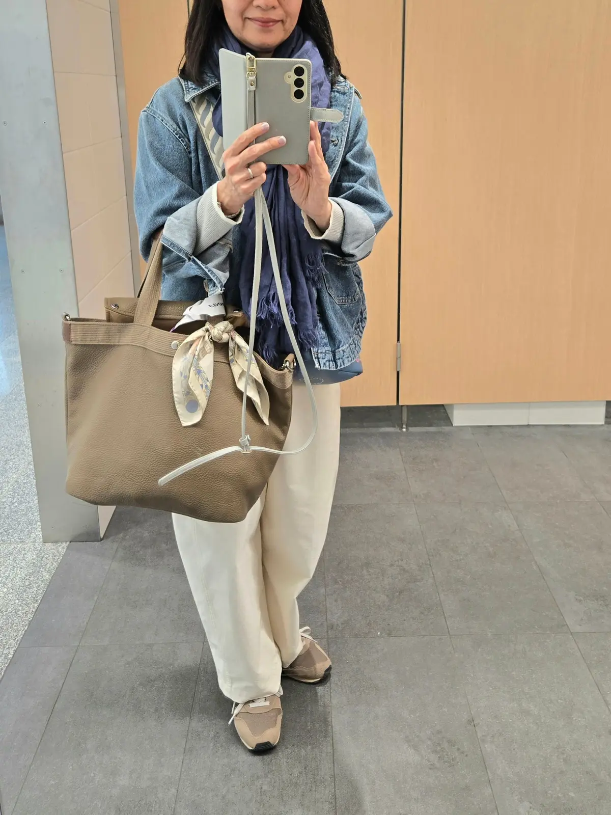 ベージュのハンドバッグを持つ女性。デニムジャケットに白いパンツ、スニーカーを着用。首にはブルーのスカーフを巻いている。スマートフォンで写真を撮っている。
