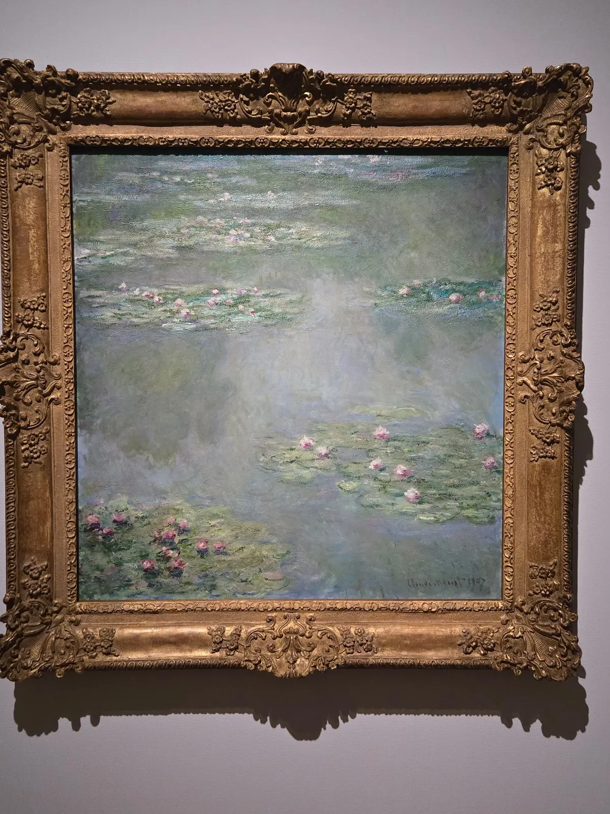 印象派の画家クロード・モネの絵画「睡蓮」。水面にピンク色の睡蓮が浮かび、緑と水色の色彩が広がる。金色の装飾的な額縁で飾られている。