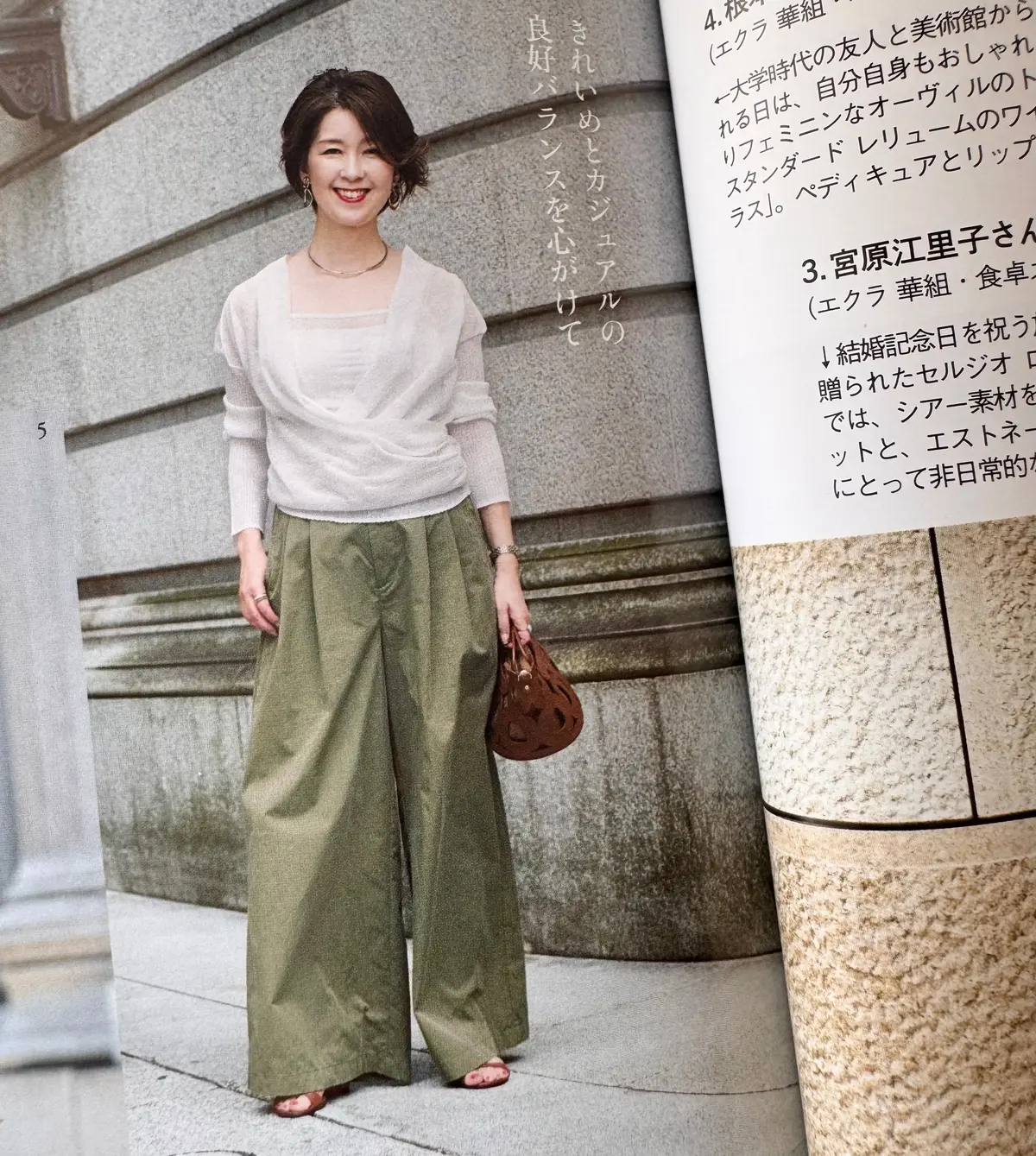 ベージュのニットに、オリーブグリーンのワイドパンツを合わせた女性。右手にリングをつけ、上品な装い。背景には雑誌のページが見える。